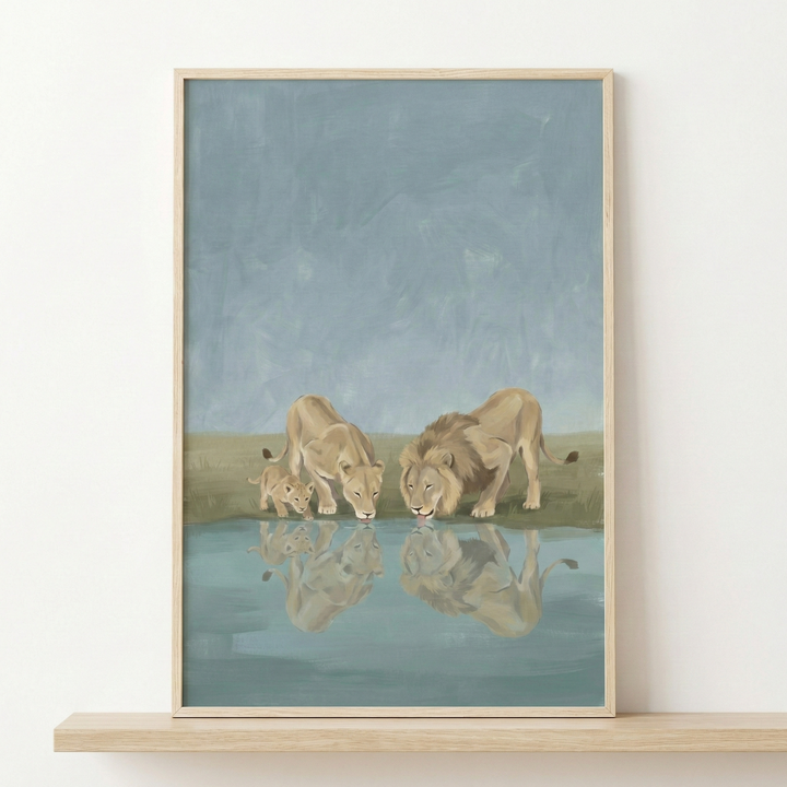 A framed piece of artwork depicting a family of lions at a watering hole. The illustration features a male lion with a mane, a female lioness, and a small cub, all leaning down to drink from calm water. Their forms are mirrored in the reflection below. The style is soft and textured, using a muted color palette of tans, light greens, and dusty blues. The artwork is enclosed in a simple, light-colored wooden frame and is shown resting on a thin wooden shelf against a plain white background.