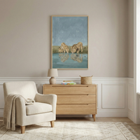 A serene room features a large vertical framed art piece depicting a male lion, a female lion, and a small cub drinking at a watering hole, with their reflections mirrored in the water. Below the art, a light oak dresser holds a small table lamp, books, and a woven basket. To the left, an upholstered armchair has a cozy textured blanket draped over it. The room has tauve walls with white wainscoting and sits on a light-colored patterned rug over wood floors.