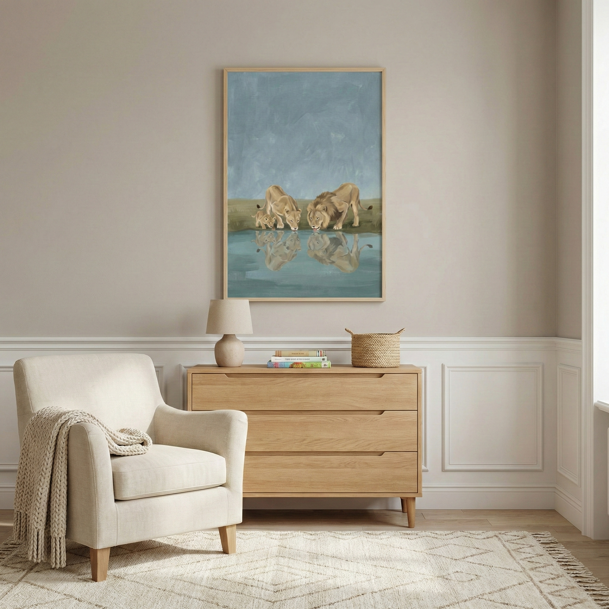 A serene room features a large vertical framed art piece depicting a male lion, a female lion, and a small cub drinking at a watering hole, with their reflections mirrored in the water. Below the art, a light oak dresser holds a small table lamp, books, and a woven basket. To the left, an upholstered armchair has a cozy textured blanket draped over it. The room has tauve walls with white wainscoting and sits on a light-colored patterned rug over wood floors.
