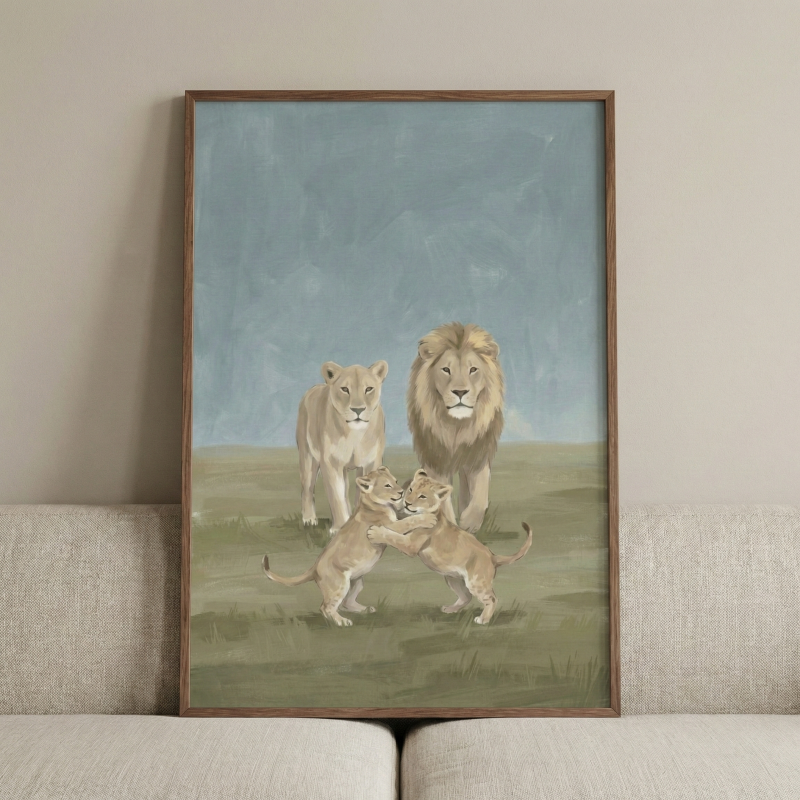 An illustration in a thin wooden frame depicts a family of lions in a grassy field. A male lion with a full mane and a lioness stand together in the background, while two lion cubs embrace in the foreground. The art features a muted color palette of tan, green, and blue-gray, with visible brushstrokes creating a soft, textured appearance. The framed work is hung on a neutral-toned wall above a light-colored fabric sofa.