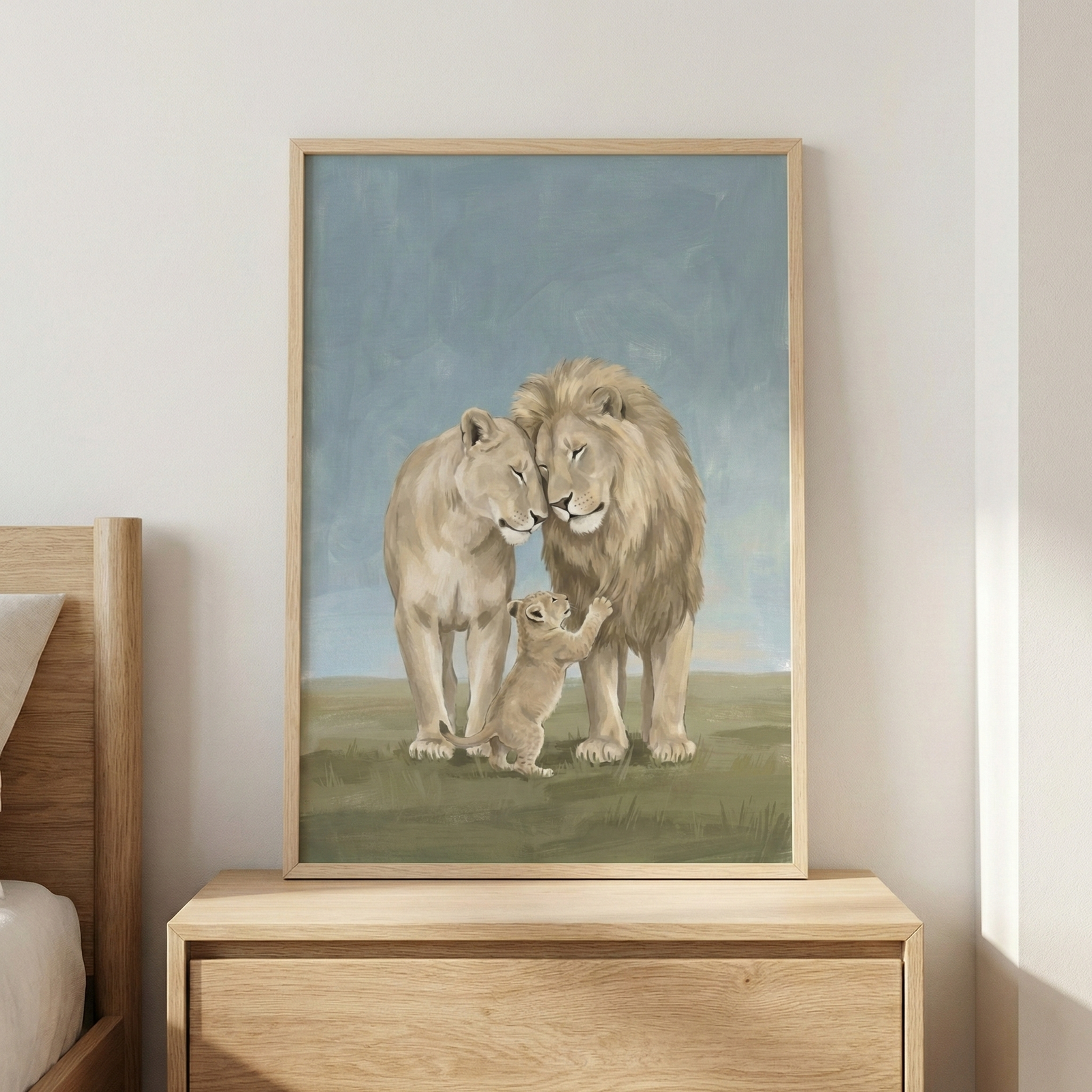 A framed piece of wall art above a wooden dresser featuring a gentle illustration of a lion family. A male lion with a full mane and a lioness lean their heads together in a loving pose. A small cub is nestled between them, reaching up towards the male lion. The artwork uses a soft color palette of beige, tan, and muted green against a hazy blue background. The image is set in a light wood frame on a neutral wall next to a bed.