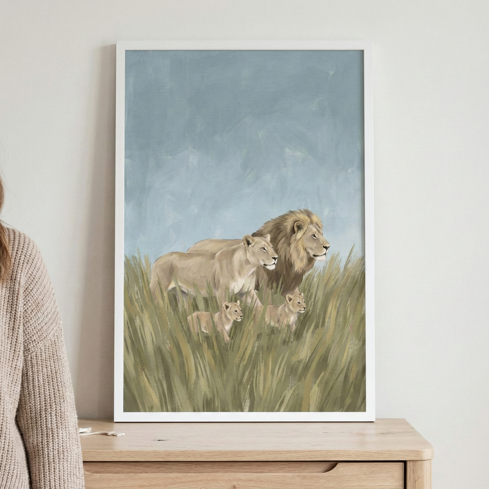 A vertical framed artwork depicts a family of lions walking through a field of tall grass. The scene includes a large male lion with a full mane, a lioness, and two small cubs. They are shown in profile, moving across the lower half of the frame. The grass is rendered in shades of green and gold, while the background consists of a soft, textured sky in light blue. The illustration uses a muted, earthy color palette and features soft, visible strokes throughout.