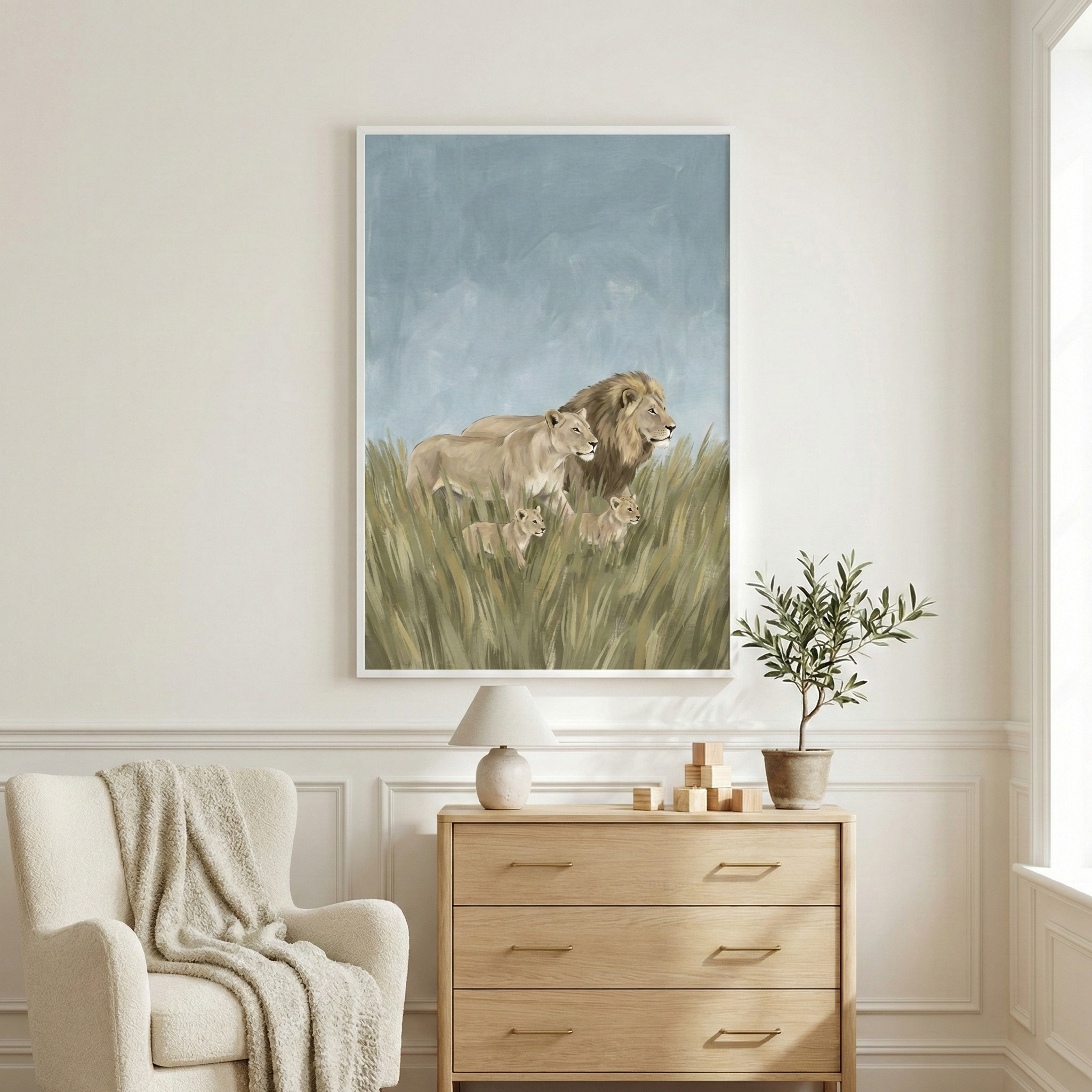 A vertical framed artwork hangs on a white wall above a light wood three-drawer dresser. The art depicts a lion family—a male, a female, and two small cubs—walking through tall grass under a hazy blue sky. On the dresser sits a simple white lamp and small wooden blocks. To the right is a green potted plant. In the foreground to the left, a cream-colored armchair is partially visible, draped with a textured knit throw blanket, creating a neutral and serene room setting.