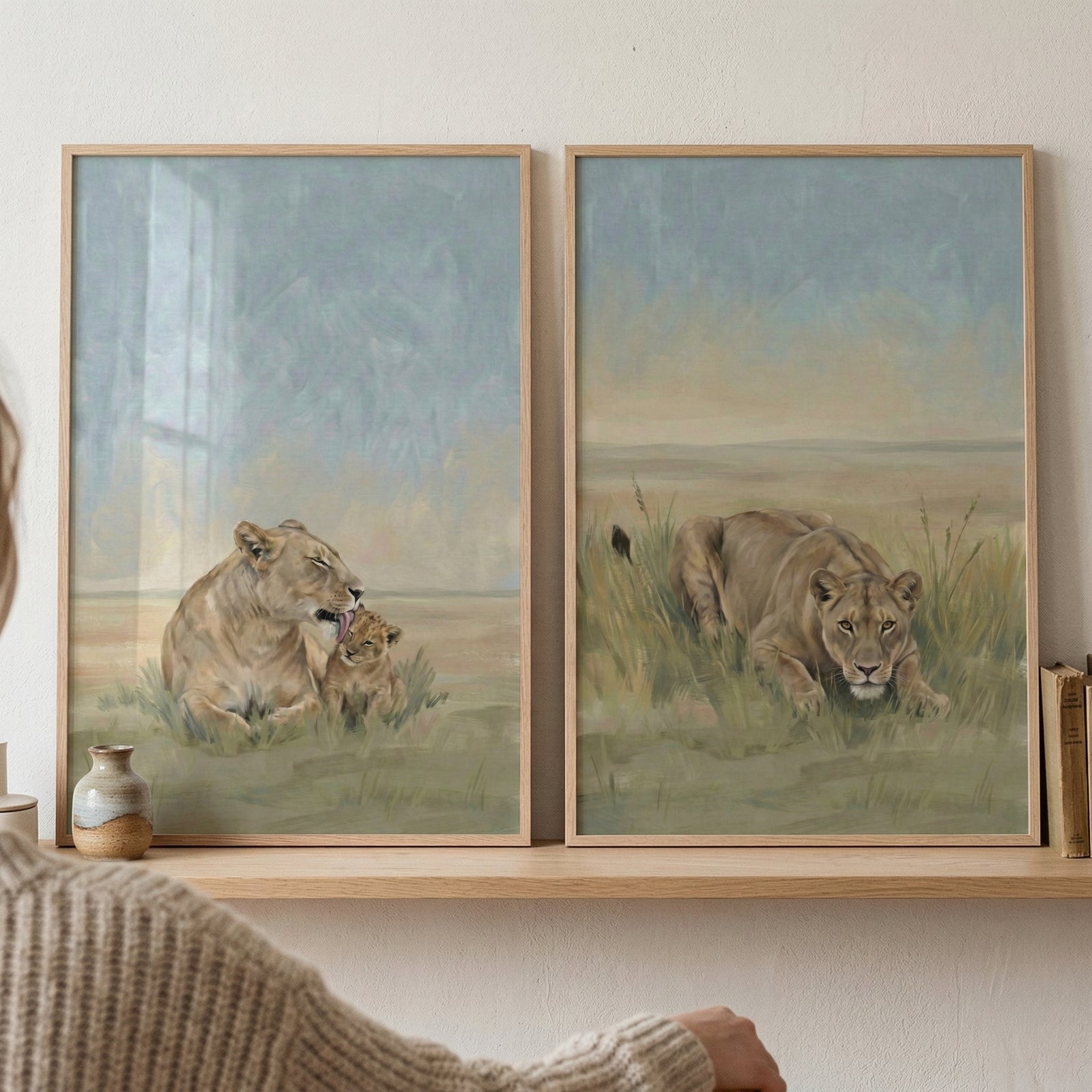 Two framed paintings show lions resting in a soft grassy savanna landscape. One artwork shows a lioness gently grooming a small cub while the other shows a lioness crouched low in the grass as if stalking prey. The warm muted tones create a calm natural wildlife scene.
