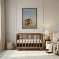 A neutral-toned nursery features a warm wood crib centered against a wall with elegant panel molding. Above the crib hangs a large framed artwork depicting a majestic lion in a grassy landscape under a soft sky. To the right, a cream rocking chair with a chunky knit blanket sits next to a small side table and a lamp. On the left, a woven basket holds soft toys. A light-colored, textured rug with fringed edges covers the floor, completing the calm and inviting space.