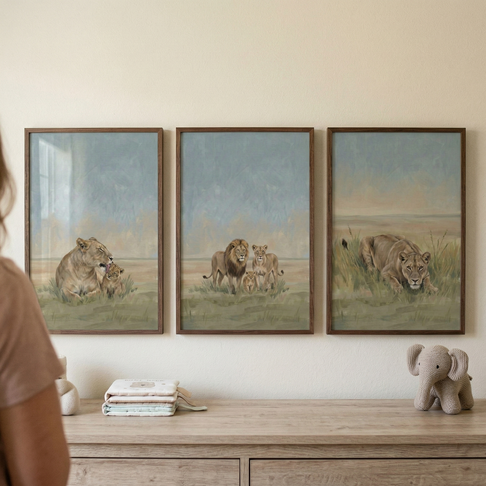 Three framed lion-themed illustrations hang on a neutral wall above a wooden dresser. Each frame contains a soft, muted-tone scene of wildlife in a grassy field. The left image shows a lioness with her cub. The middle one features a lion family including a male, a female, and two cubs. The right image depicts a lioness crouched in tall grass. On the dresser below, there is a stack of folded linens and a small, gray knitted toy elephant. A person's shoulder is visible in the left foreground.
