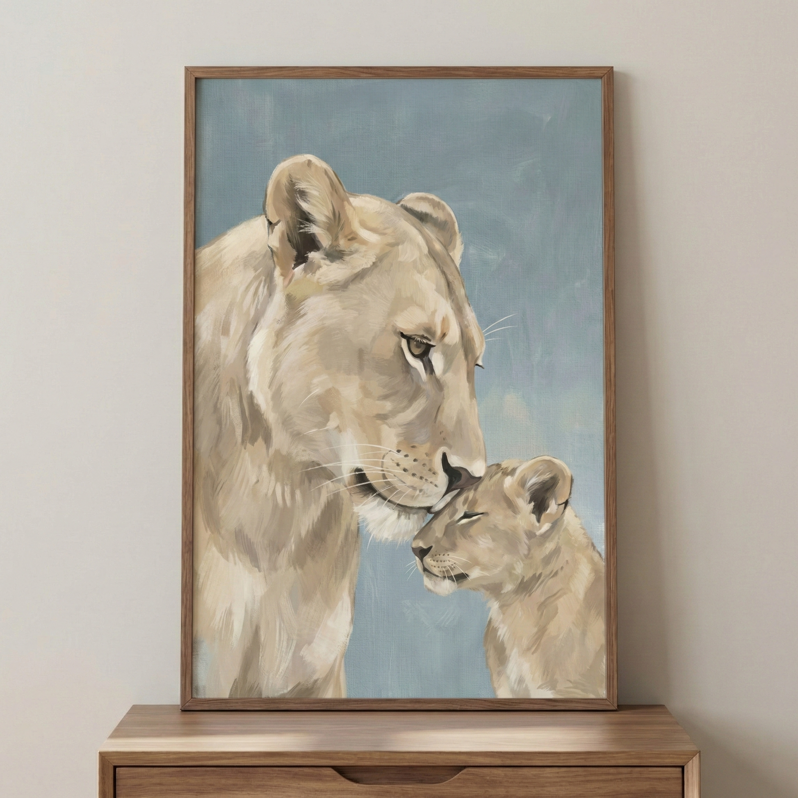 A vertical framed artwork captures a mother lion gently nuzzling her cub. The large lioness leans down, her nose softly touching the forehead of the cub, who has its eyes closed in a moment of peace. The illustration features visible, layered strokes in a palette of cream, sand, and light brown. The background is a flat, muted blue-gray. The piece is housed in a simple wooden frame and displayed on a light brown wooden tabletop against a plain, off-white wall.