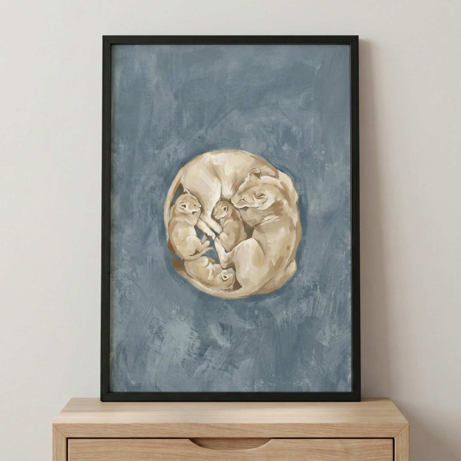 A vertical black-framed piece of art sits on a light wooden dresser. The artwork features a group of big cats curled together into a circular formation in the center. A larger tan lioness is nestled with three smaller cubs, all rendered with textured brushstrokes in shades of beige and cream. The background is a solid, muted blue with visible texture and variations in tone. The overall composition creates a sense of warmth and protection within the cool-toned surrounding space.