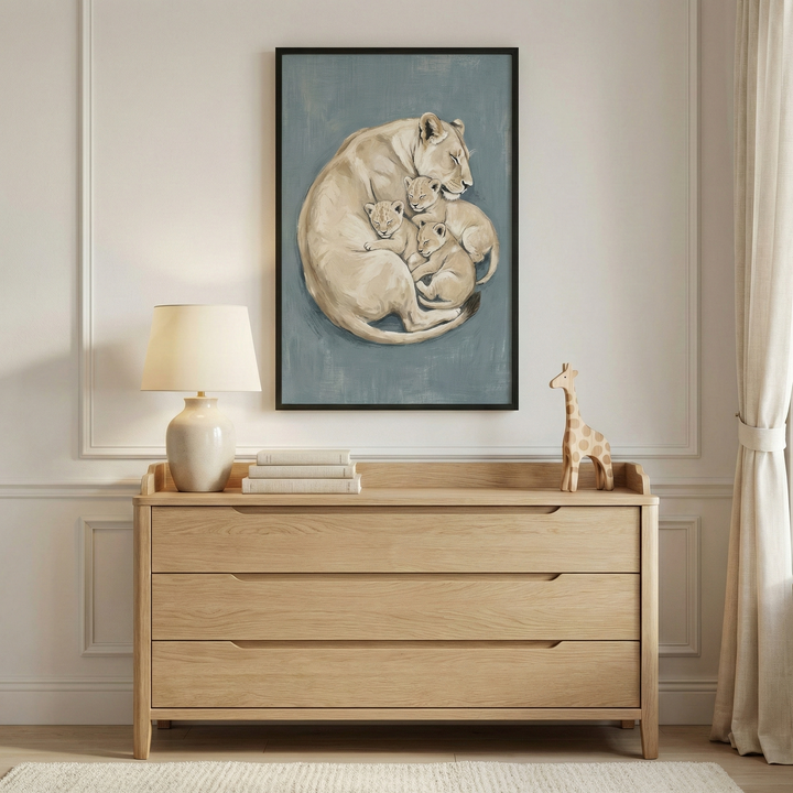 This image captures a serene and modern nursery or bedroom setup. Centered on a wall with elegant, light-colored picture frame molding is a large rectangular framed art piece. The artwork depicts a tender scene of a mother lioness and her three small cubs curled together in a close, circular embrace. The subjects are rendered in soft, warm beige tones that contrast with the muted blue-gray background of the print. The frame itself is thin and black.
