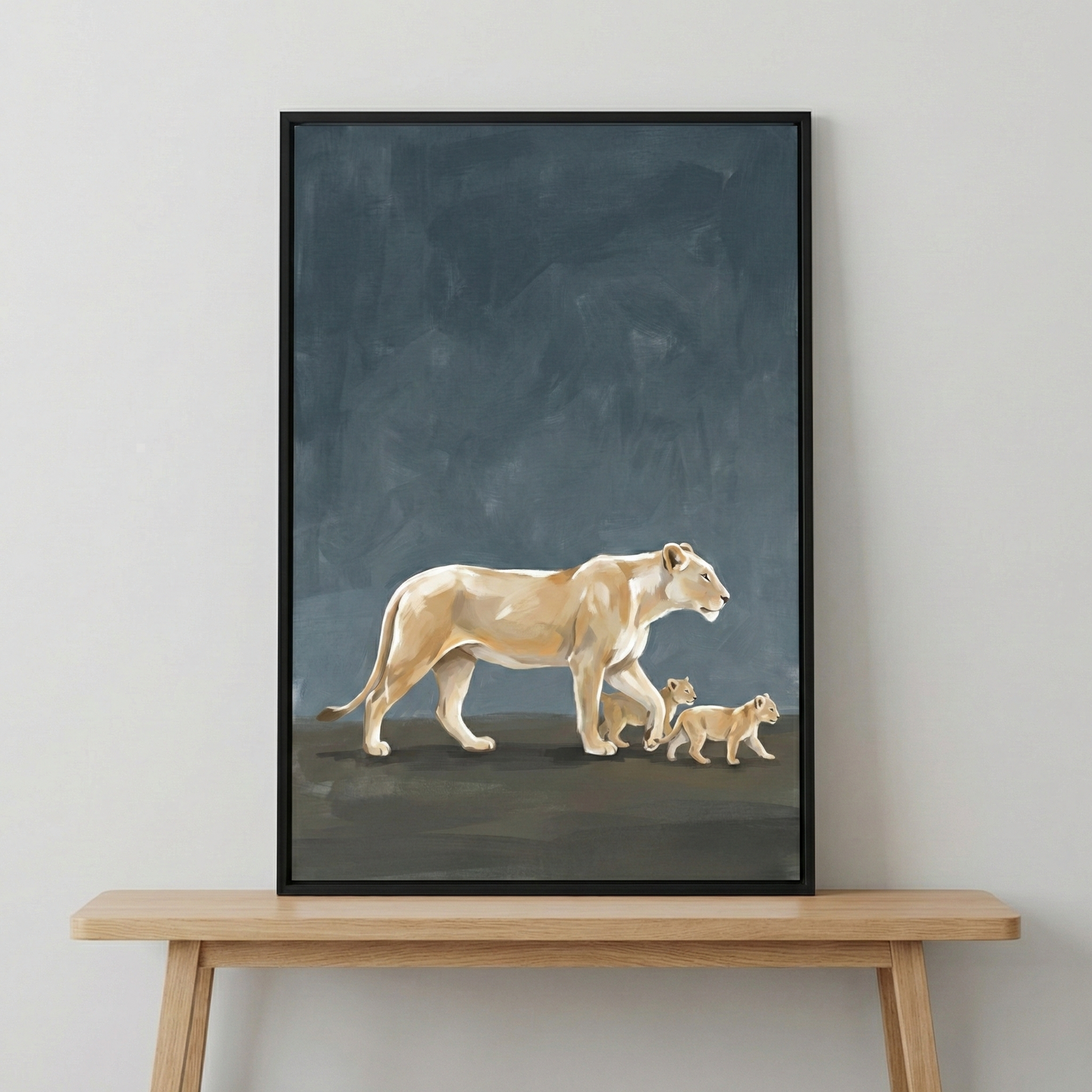 A vertical piece of wall art in a slim black frame sits on a light wood console table against a plain, light-colored wall. The artwork features a large female lion walking across a dark landscape, followed closely by two small cubs. The style of the art is expressive and minimalist, with visible brushstrokes and a palette of light cream and tan for the animals. The background of the art is a deep, textured navy blue that contrasts with the lighter subjects. 