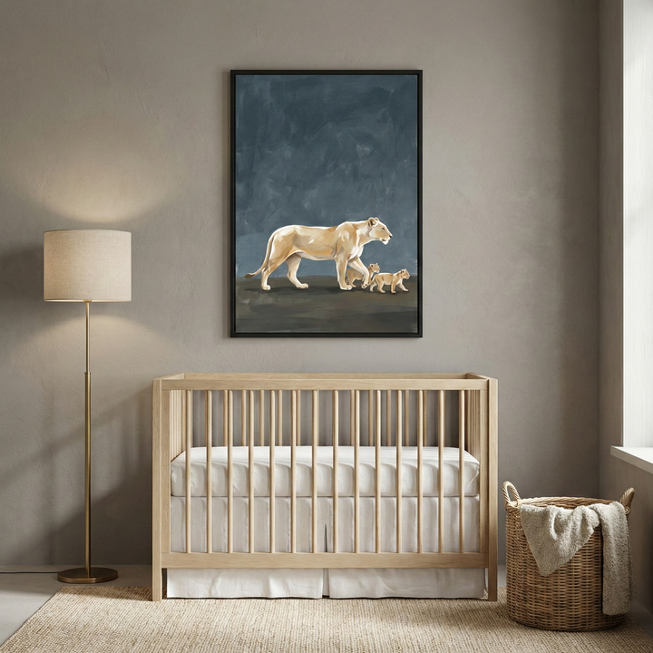 A serene and modern nursery features a large framed artwork as its centerpiece on a soft taupe wall. The illustration depicts a light-colored lioness followed by two small cubs walking across a dark, textured slate-blue background. Positioned directly beneath the artwork is a light-toned wooden crib with a simple slatted design, dressed in crisp white bedding. To the left of the crib stands a minimalist floor lamp with a slender metallic base and a neutral drum shade. 