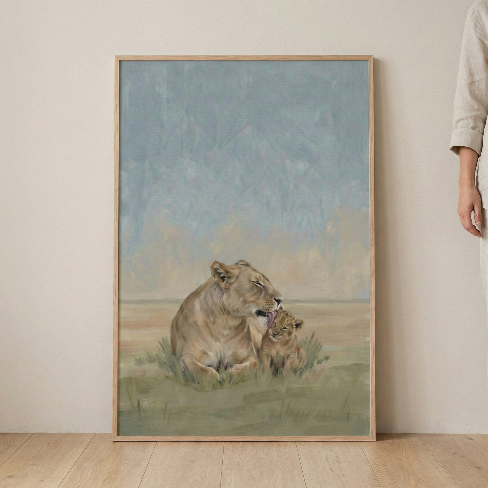 A vertically oriented artwork in a light-colored wooden frame depicts a tender moment between a lioness and her cub. The lioness, with a soft beige and tan coat, sits nestled in green grass and gently licks the top of her cub's head. The cub sits beside its mother, its eyes closed in a peaceful expression. The background is an open landscape with a hazy blue sky and muted golden horizon. The style features visible brushwork and a soft color palette, creating a gentle scene of nature.