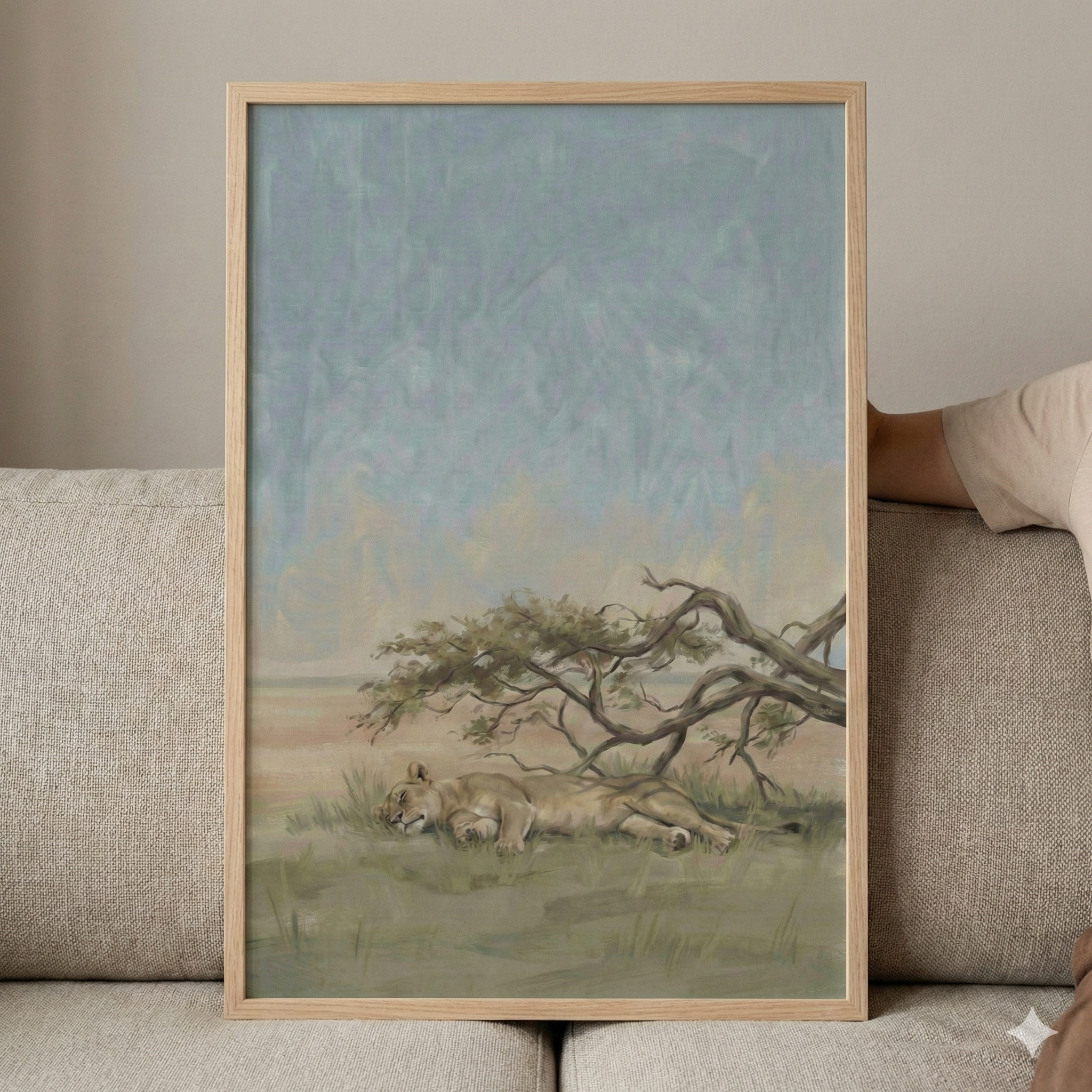 A vertical artwork in a light wooden frame is propped against a grey fabric sofa. The illustration depicts a peaceful savanna scene where a lioness sleeps on the grass beneath the shade of a weathered, gnarled tree. The landscape is rendered in muted earthy tones of tan and soft green. Above, a vast, hazy sky is filled with blended strokes of pale blue and light yellow, creating a calm atmosphere. A portion of a person's arm in a beige sleeve is visible on the right side of the frame.