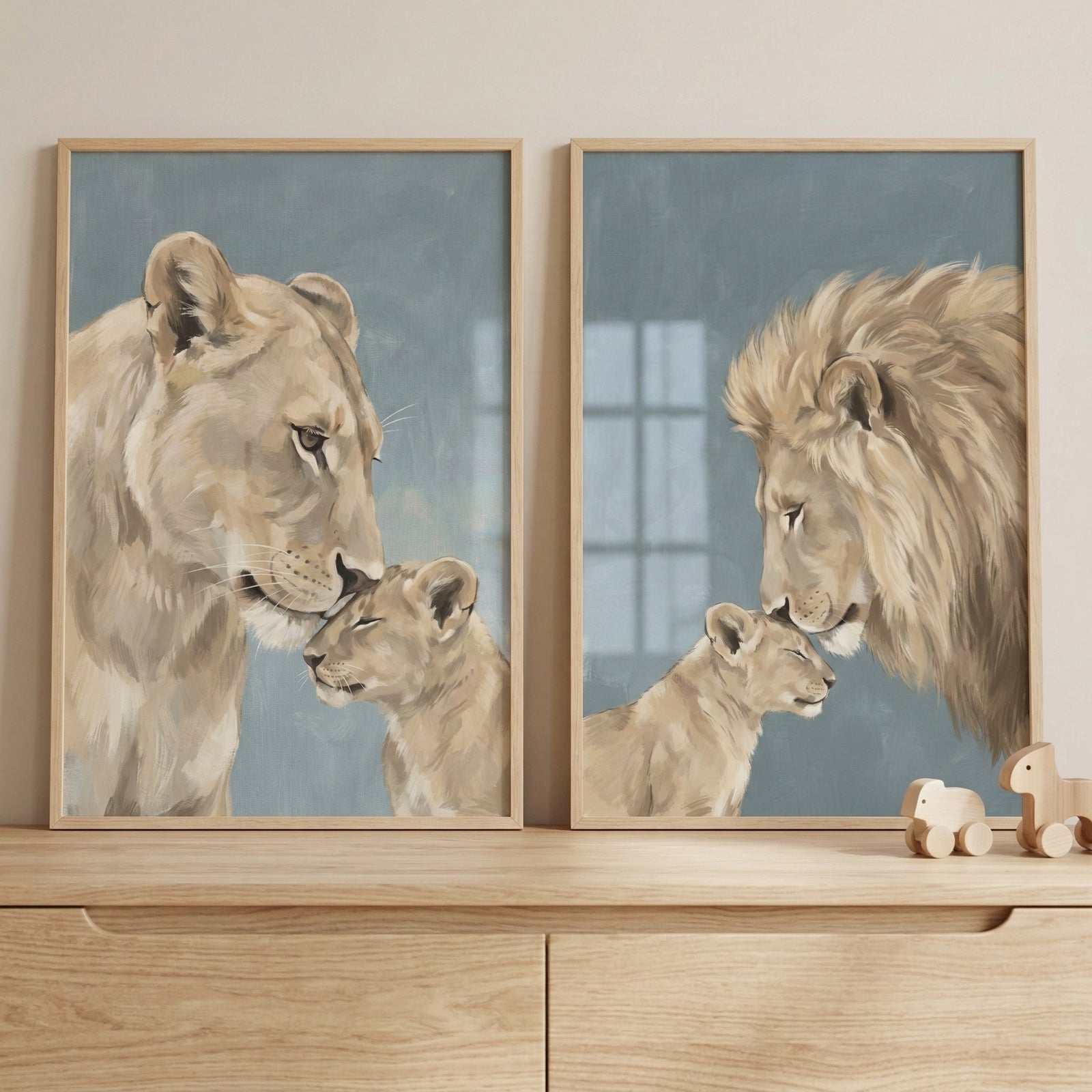 Two vertical wooden-framed art prints are displayed side-by-side on a light-colored wall above a wooden chest of drawers. The illustration on the left shows a large lioness gently nuzzling the head of a small lion cub. The illustration on the right features a large male lion with a full mane nuzzling a similar cub. Both pieces are rendered in soft beige and tan tones against a muted, dusty blue background with visible textured brushwork. To the right of the frames on the dresser sit two small, minimalist wo