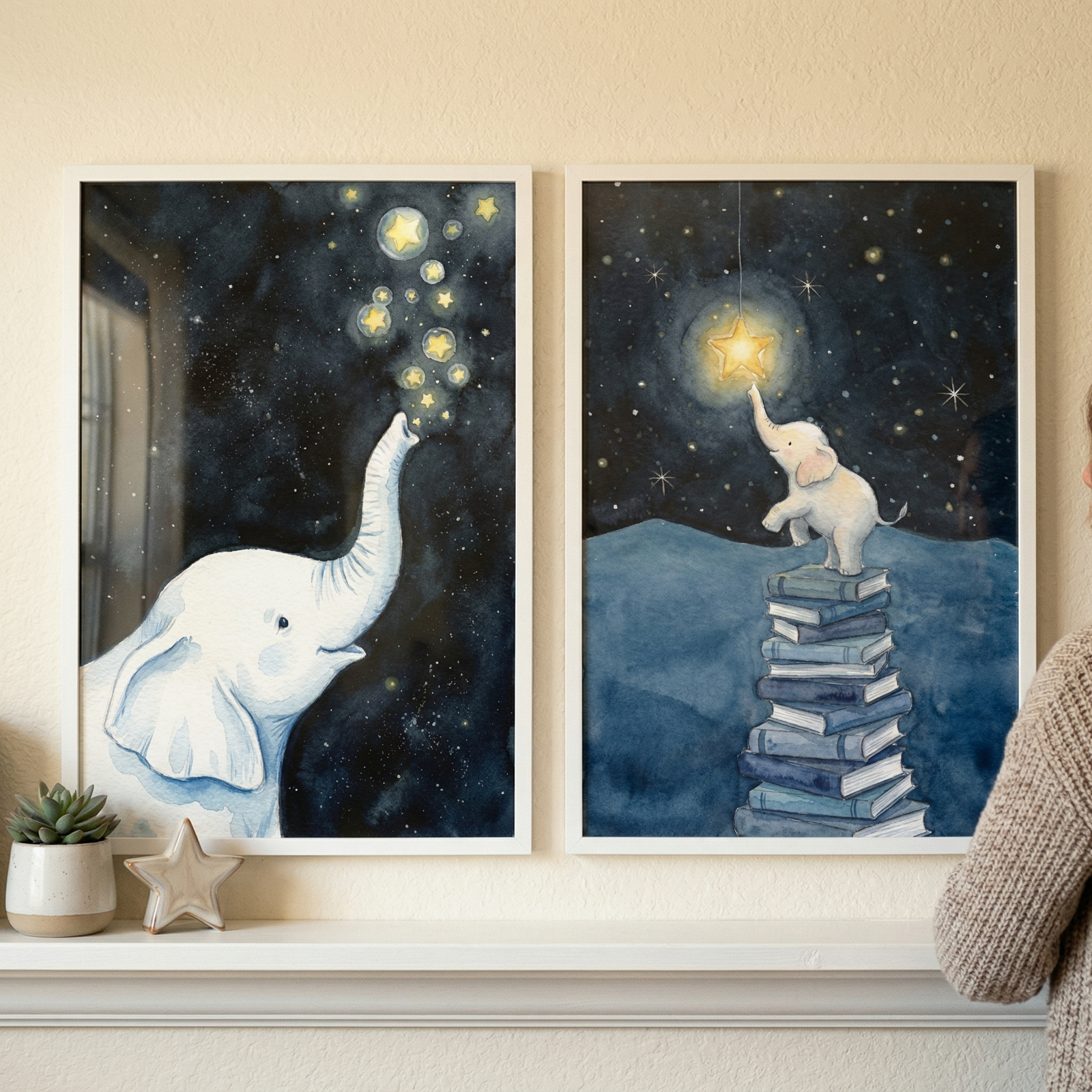 Two framed whimsical watercolor illustrations featuring a white elephant are displayed on a white mantel. The artwork on the left shows a large white elephant looking upward, its trunk raised towards a dark blue night sky filled with glowing stars. The artwork on the right depicts a smaller white elephant standing on a tall stack of blue books, reaching up to touch a bright star hanging from a string. A small potted succulent and a wooden star ornament sit on the mantel near the frames.