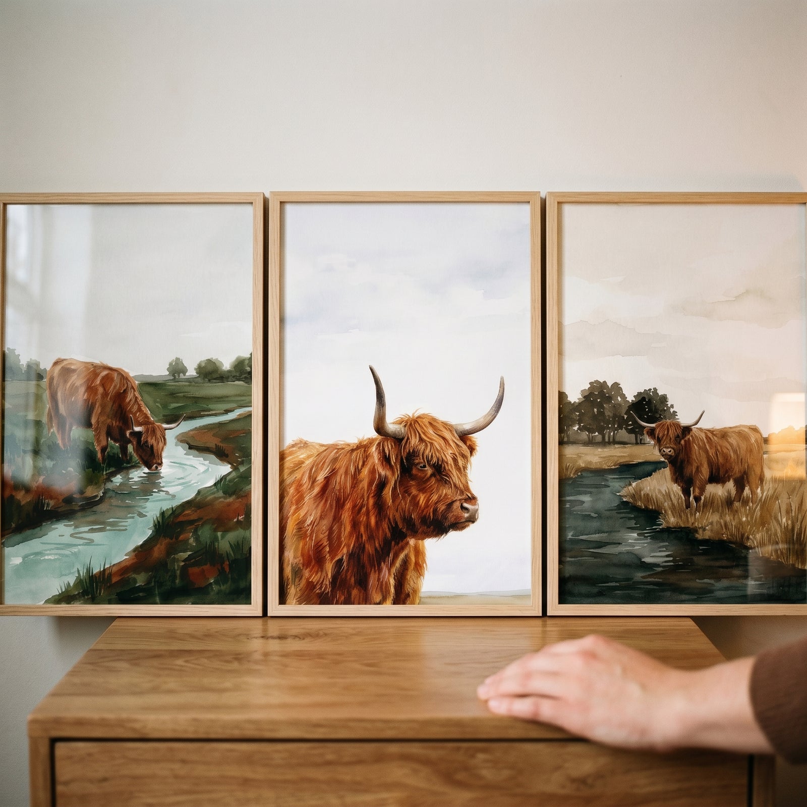 Long horned highland cow wall art set of 3 depicts cows near water and in portrait, in wooden frames on a cabinet.