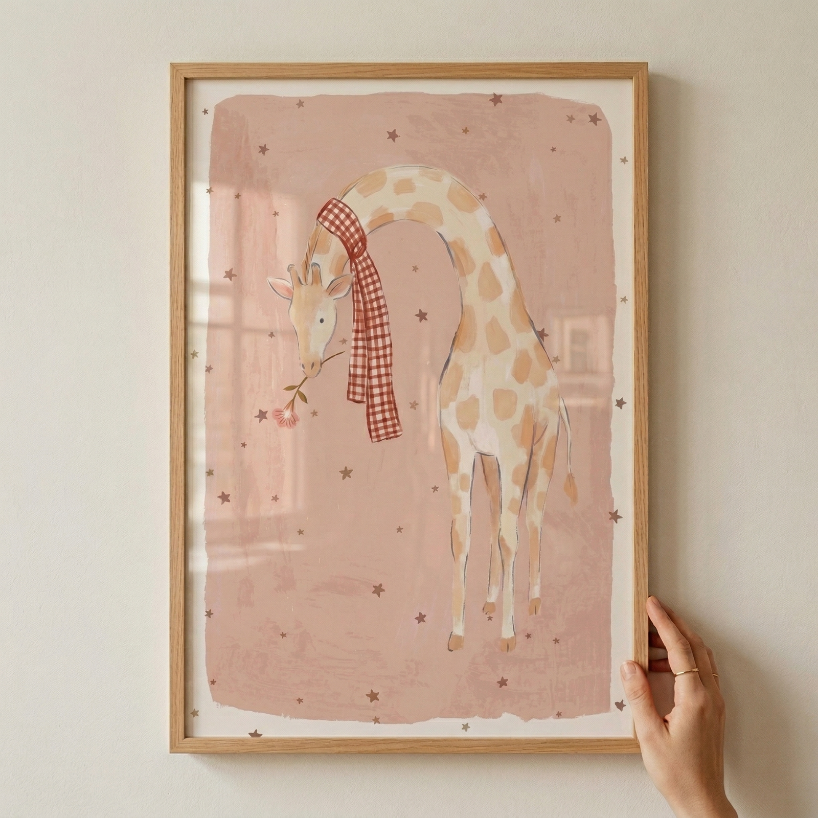 A framed illustration depicts a whimsical giraffe with its head gently bowed. The animal has soft, light spots and wears a red and white checkered scarf around its neck, while holding a small pink flower in its mouth. The background is a muted, dusty pink color with a hand-drawn textured edge, featuring a scattered pattern of tiny dark stars. The artwork is set within a light wood frame, which is being held by a hand against a neutral-colored wall.