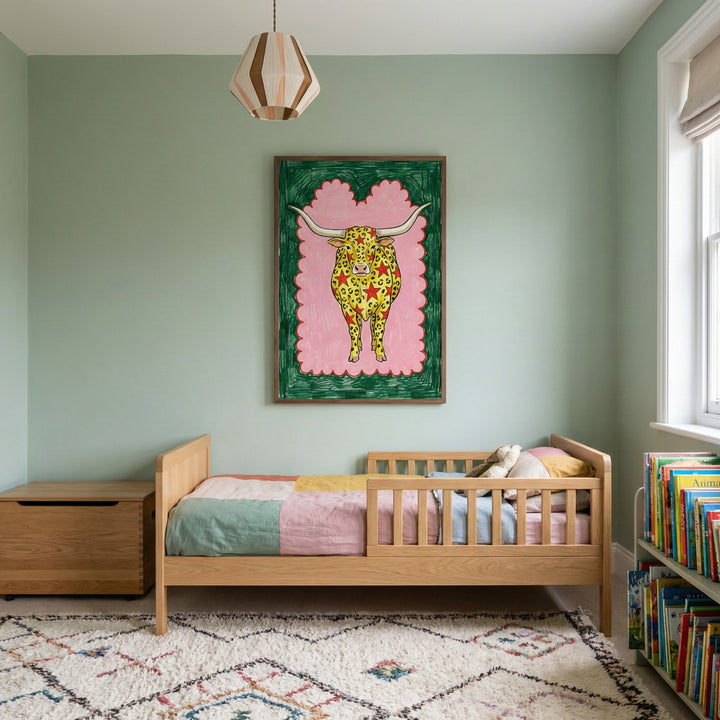 Framed whimsical artwork of a longhorn cow displayed above a child's bed in a pastel-colored bedroom with wooden furniture and a cozy rug.
