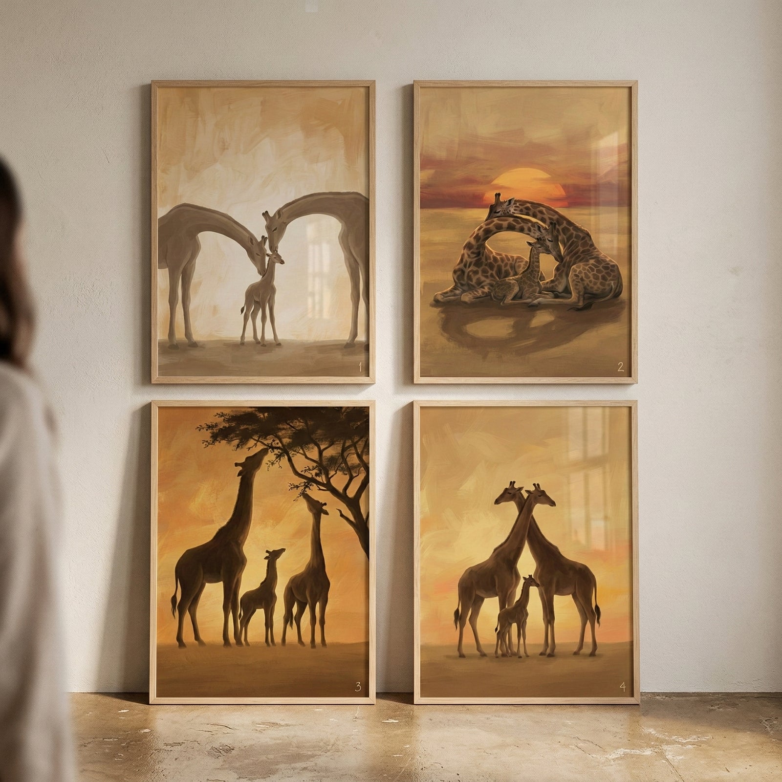 Four framed giraffe paintings arranged in a grid show warm sunset family scenes in soft earthy tones. One shows two adult giraffes bending their long necks toward a small calf standing between them, while another shows two giraffes curled protectively around a resting calf beneath a glowing sun. The other artworks show a giraffe family standing beneath a tree and two adults standing close together with a calf between them.