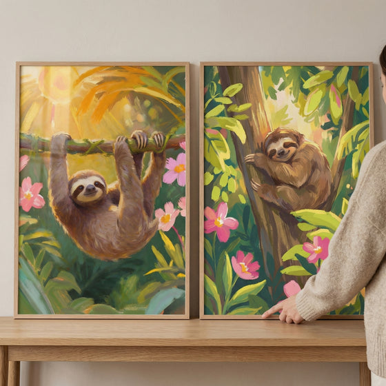 Two framed jungle paintings rest on a wooden surface while a person points at one of them. One shows a sloth hanging upside down from a branch in warm sunlight surrounded by pink flowers while the other shows a sloth curled up on a tree trunk among green leaves in a lush forest.