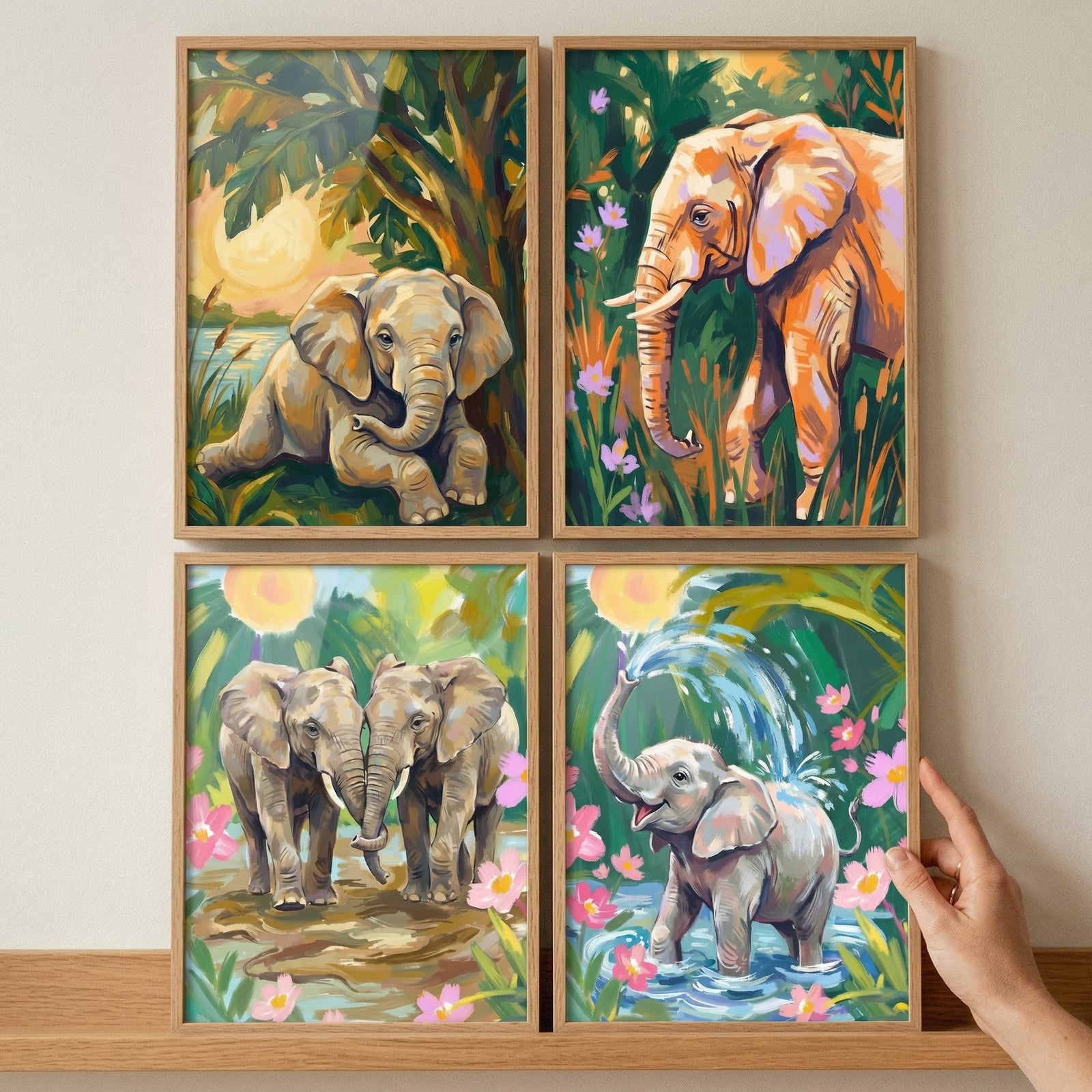A collection of four framed illustrations featuring elephants in vibrant tropical environments. The artworks are displayed in a two-by-two grid on a wooden surface. Scenes include a baby elephant lounging under leaves, an adult elephant trekking through flora, two elephants near water, and a young elephant splashing in a pond. The style is characterized by bold brushwork and a rich palette of greens, oranges, and pinks. The frames are made of light-colored wood, and a hand is visible touching one.
