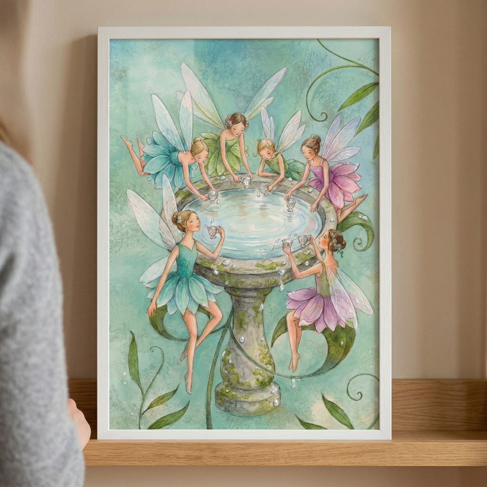 A framed vertical illustration features six delicate, winged fairies gathered around a moss-covered stone font or birdbath filled with shimmering water. The artwork is rendered in a soft, ethereal style with a palette of light blues, greens, and purples. Each fairy has translucent, insect-like wings and wears a dress reminiscent of layered flower petals in shades of teal, emerald, and lavender. 
