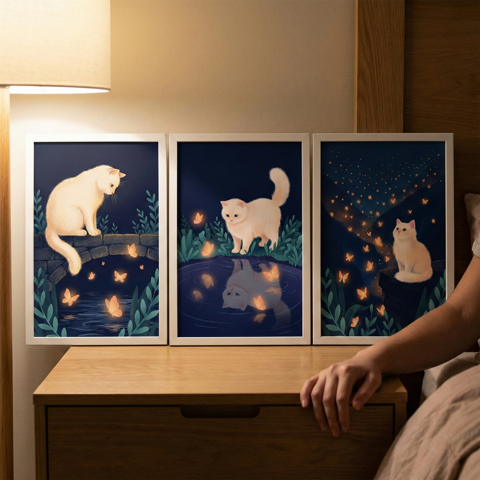 Three framed illustrative prints of a white fluffy cat sit on a wooden nightstand. Each piece shows a whimsical nighttime scene with glowing orange butterflies. The left print shows the cat on a stone bridge. The center print shows the cat by a dark pond with its reflection visible. The right print features the cat on a rock under a starry sky filled with fireflies. A person’s hand rests on the table, and a warm light from a nearby lamp illuminates the serene bedroom setting.