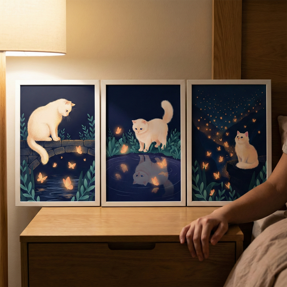 Three framed illustrative prints of a white fluffy cat sit on a wooden nightstand. Each piece shows a whimsical nighttime scene with glowing orange butterflies. The left print shows the cat on a stone bridge. The center print shows the cat by a dark pond with its reflection visible. The right print features the cat on a rock under a starry sky filled with fireflies. A person’s hand rests on the table, and a warm light from a nearby lamp illuminates the serene bedroom setting.