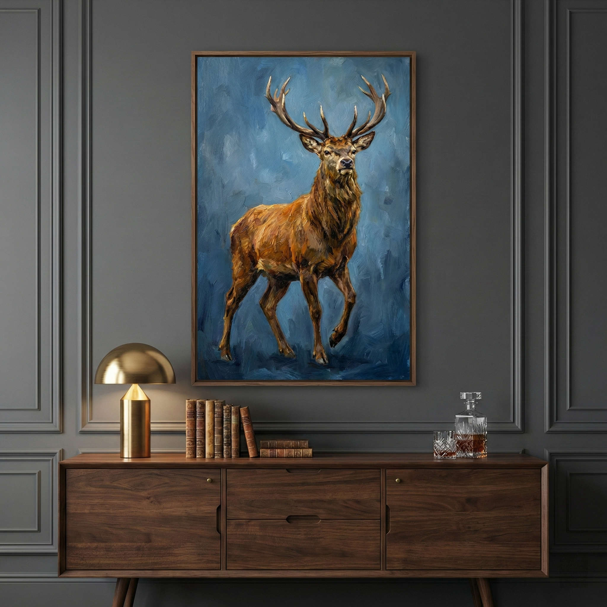 A large framed artwork of a brown stag with prominent antlers is centered on a dark grey paneled wall. The stag is rendered with thick, expressive strokes against a textured blue backdrop. Below the art piece, a dark wood mid-century style credenza holds several items: a gold mushroom-shaped lamp casting a warm glow, a row of aged books, and a glass decanter with a small glass. The room has a sophisticated, moody atmosphere with classic wall molding.