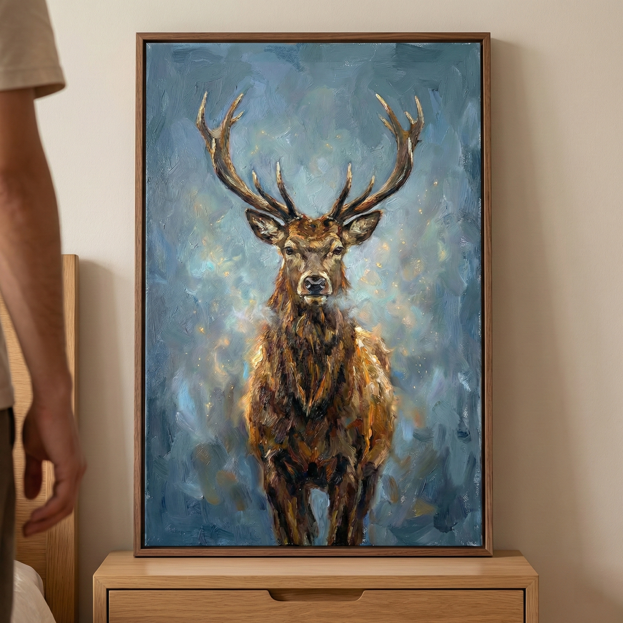An expressive, framed canvas art piece featuring a majestic stag with large, branching antlers. The artwork is created with thick, visible textured strokes in earthy shades of brown, tan, and gold, set against a mottled, atmospheric blue background with subtle glowing highlights. The piece is encased in a simple wooden frame and sits on a light-colored wooden nightstand in a room with neutral walls. To the left, a portion of a person's side and arm is visible.