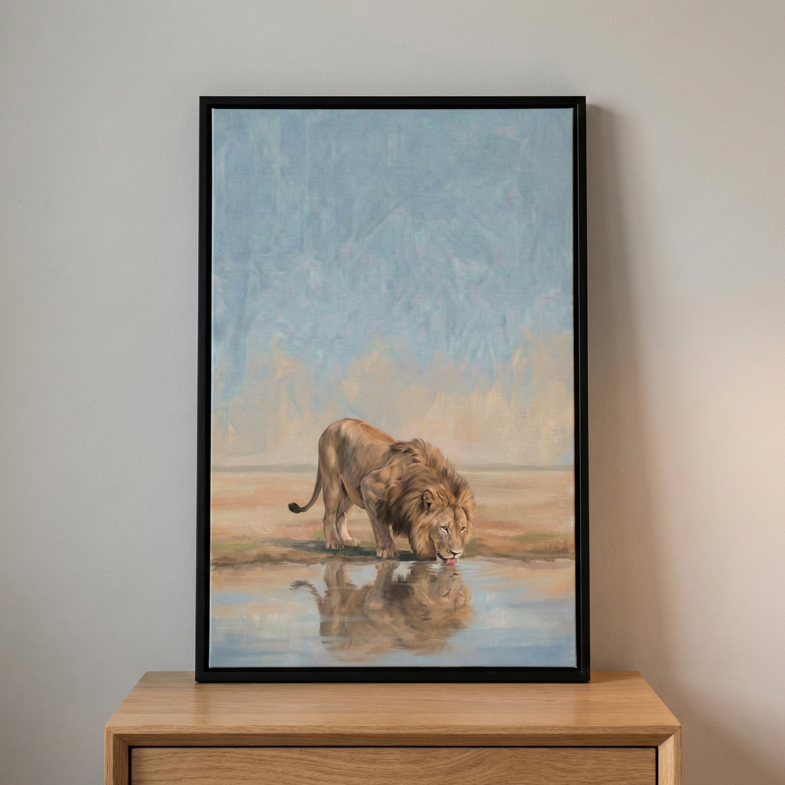 A vertically oriented artwork featuring a majestic male lion is shown in a thin black frame. The lion, with a thick, dark mane and golden-brown fur, is depicted leaning down to drink from a calm, still body of water. A clear reflection of the lion's head and body is visible in the water's surface. 

