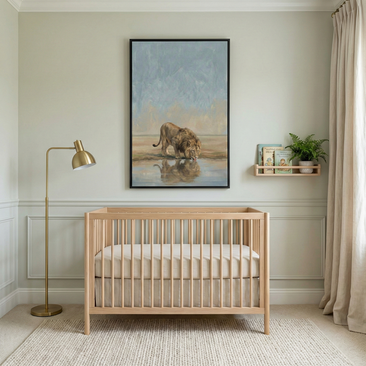 A serene and modern nursery decorated in neutral tones. A light-colored wooden crib sits in the center, dressed with simple cream bedding. Above the crib hangs a large framed piece of art featuring a realistic-style illustration of a lion drinking from a pool of water, reflected on the surface. To the left, a slim gold floor lamp stands beside the crib. On the right, a small wooden wall shelf holds books and a green plant. Textured beige rug and light curtains complete the calm atmosphere.