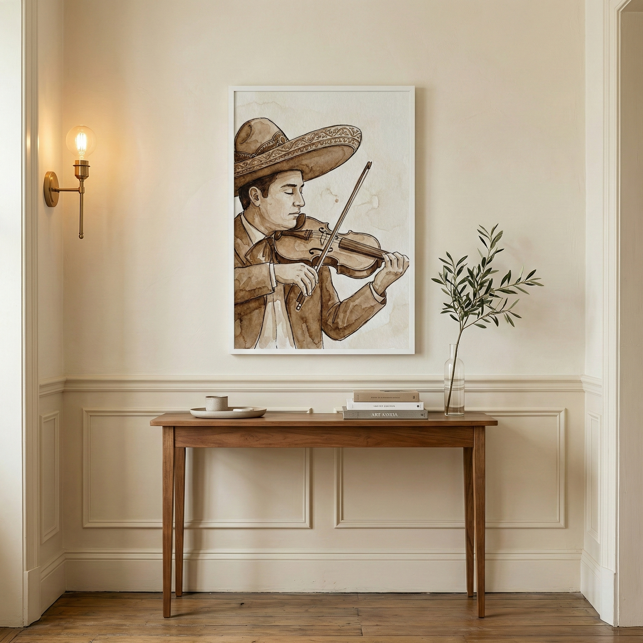 An interior setting features a framed piece of art on a cream-colored wall above a wooden console table. The artwork shows a man in a traditional wide-brimmed hat and brown suit playing a violin, rendered in warm sepia tones. The table holds a small dish, a stack of books, and a glass vase with a green leafy branch. To the left, a brass wall lamp with a glowing bulb illuminates the space. The room is detailed with elegant wall molding and light wood floors.