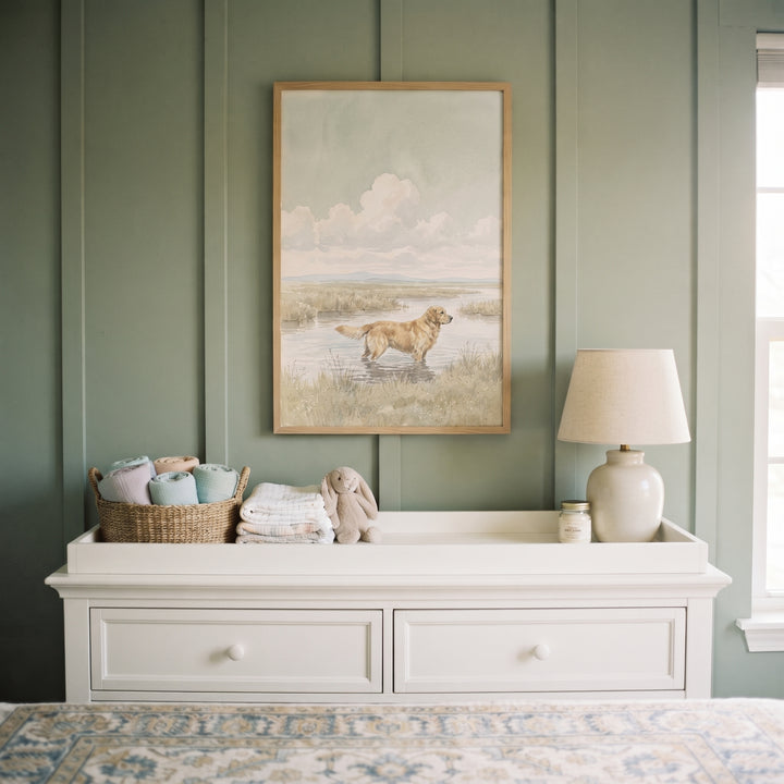 Marsh landscape golden retriever dog wall art above a white dresser against a sage green paneled wall with a lamp.