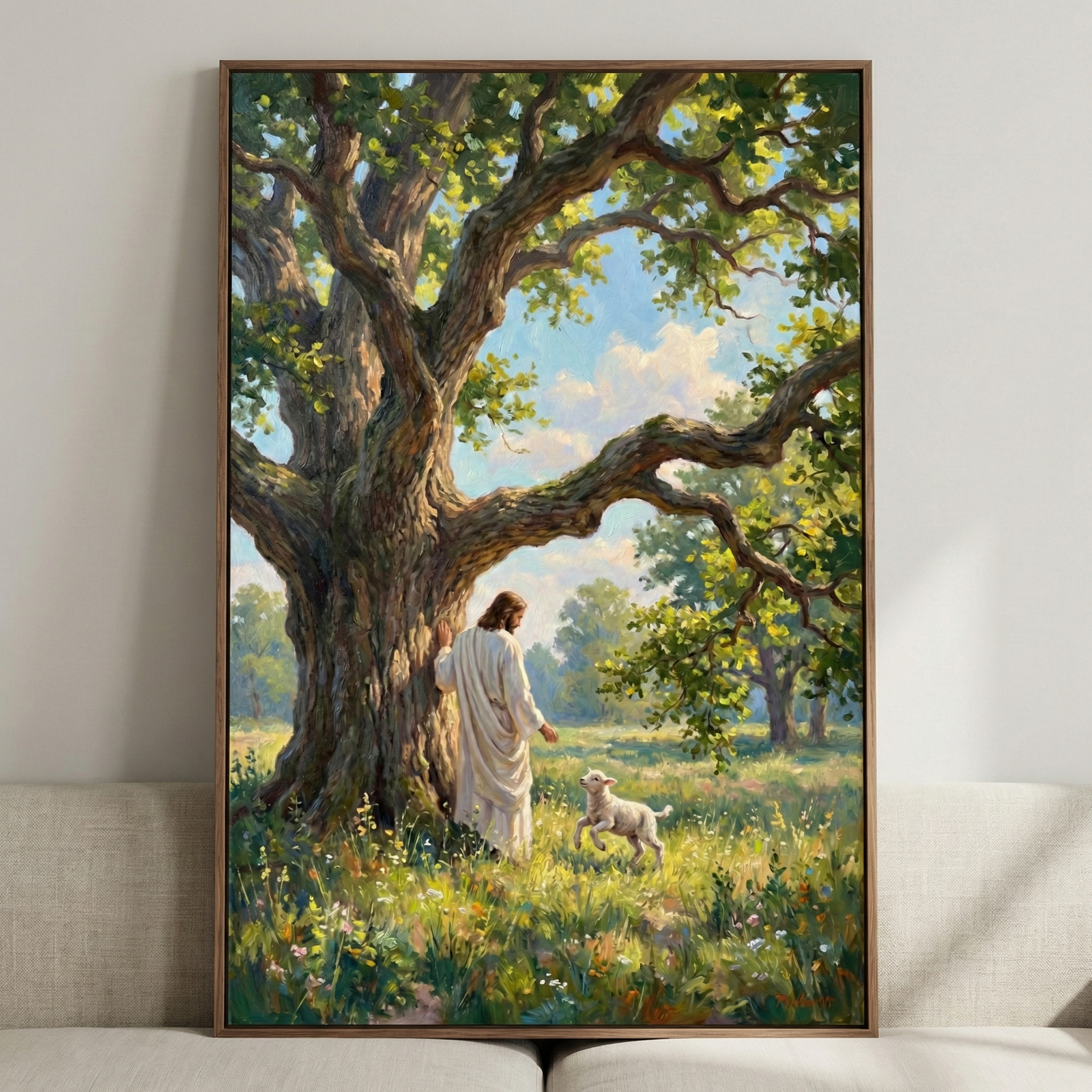 A framed vertical artwork depicts Jesus Christ in a long white robe standing beside a massive, ancient tree. He looks toward a small white lamb frolicking in a lush meadow filled with wildflowers and tall grass. Sunlight filters through the dense green canopy of the tree, creating a warm, peaceful glow. The piece uses soft, visible brushstrokes and is enclosed in a dark wood frame, hanging on a light-colored wall above a neutral-toned couch.