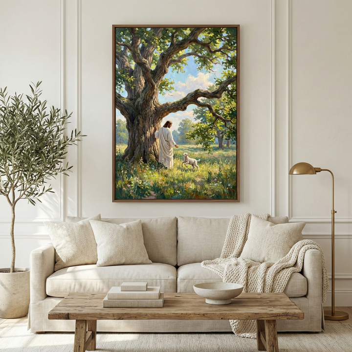 A bright living room features a minimalist design with a beige fabric sofa, plush pillows, and a textured throw blanket. Above the sofa hangs a framed artwork of Jesus Christ in a white robe, standing in a sun-drenched meadow beneath a large, leafy tree. He is looking at a small white lamb in the grass. A rustic wooden coffee table with books and a bowl sits in front of the sofa. A potted olive tree is to the left, and a gold floor lamp is on the right.