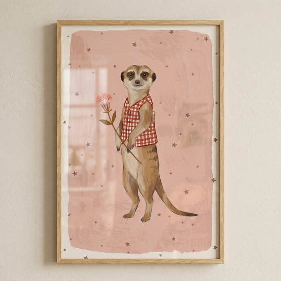 A vertical illustration of a meerkat standing upright is displayed within a light-colored wooden frame. The meerkat is depicted with soft tan and brown fur, wearing a red and white checkered vest. It holds a single, delicate pink flower with a long stem and small green leaves. The background is a textured, light pink rectangular field, adorned with several small, dark brown stars scattered around the figure. The framed artwork is shown hanging on a neutral, off-white wall with a subtle texture.