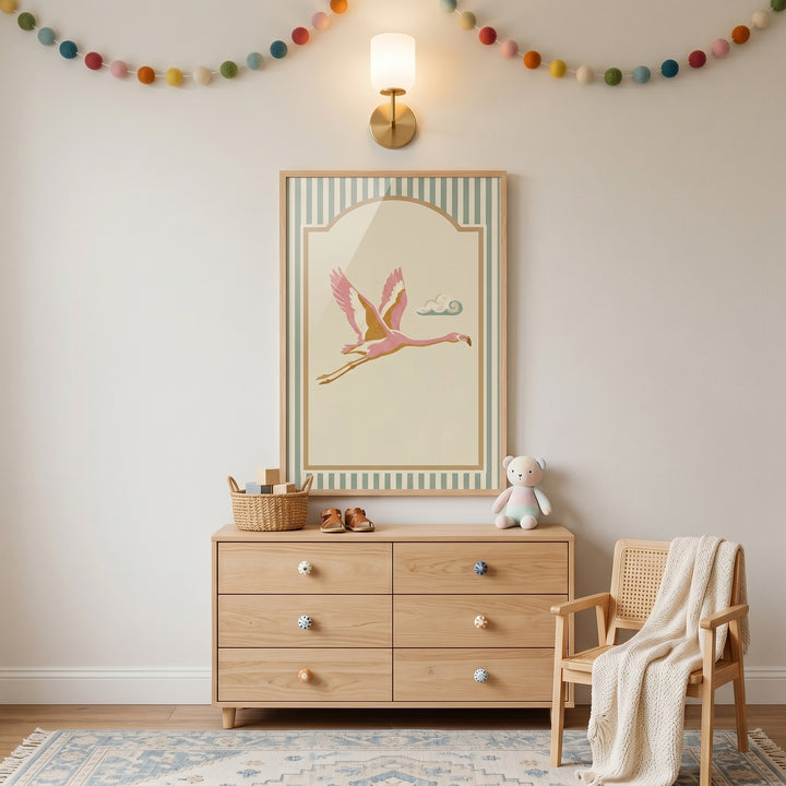 Mid century pink flamingo wall art hangs above a wooden dresser in a room with a pom-pom garland and a wooden chair.