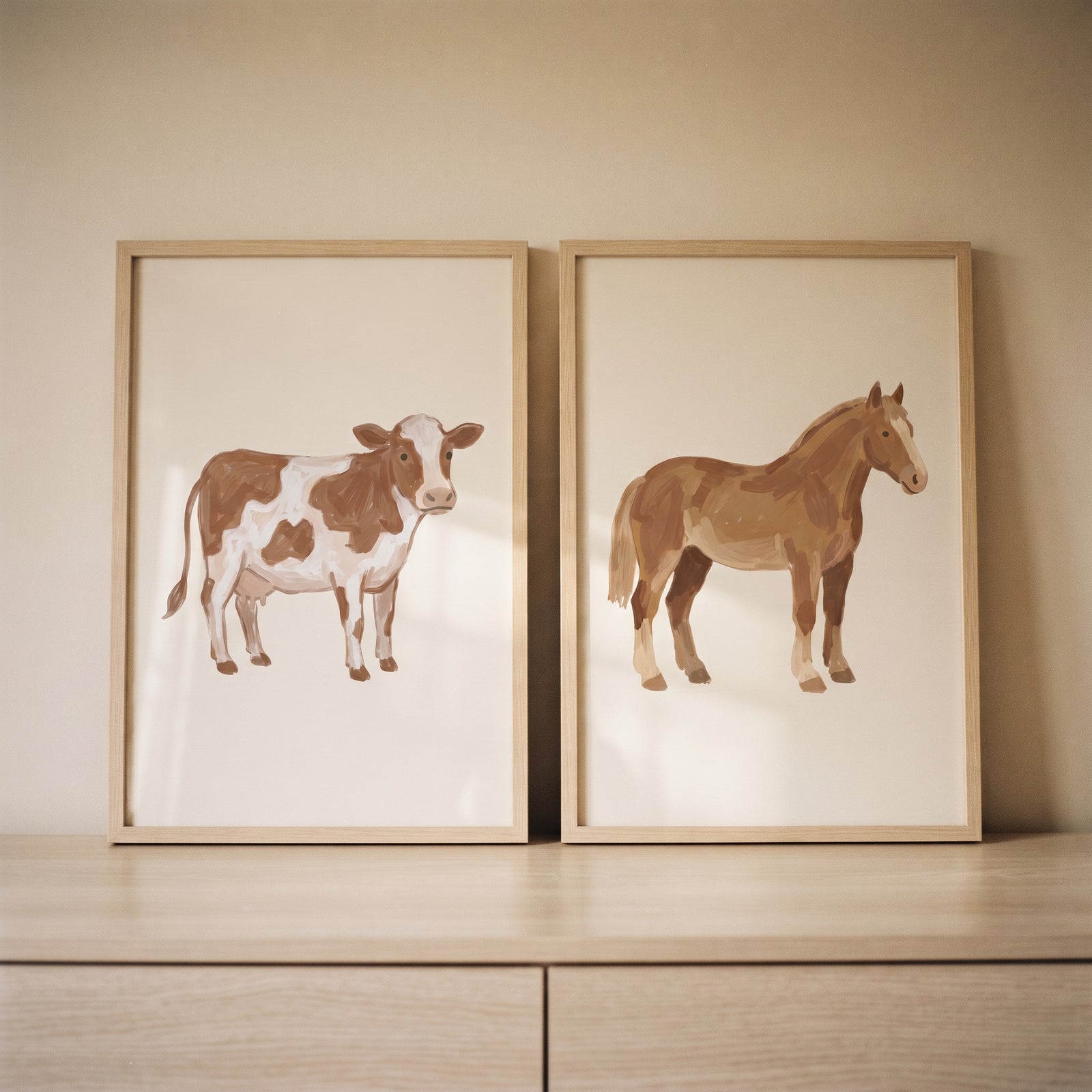 Minimalist beige farm animals wall art set of 2 featuring a spotted cow and a horse in frames on a wooden dresser.