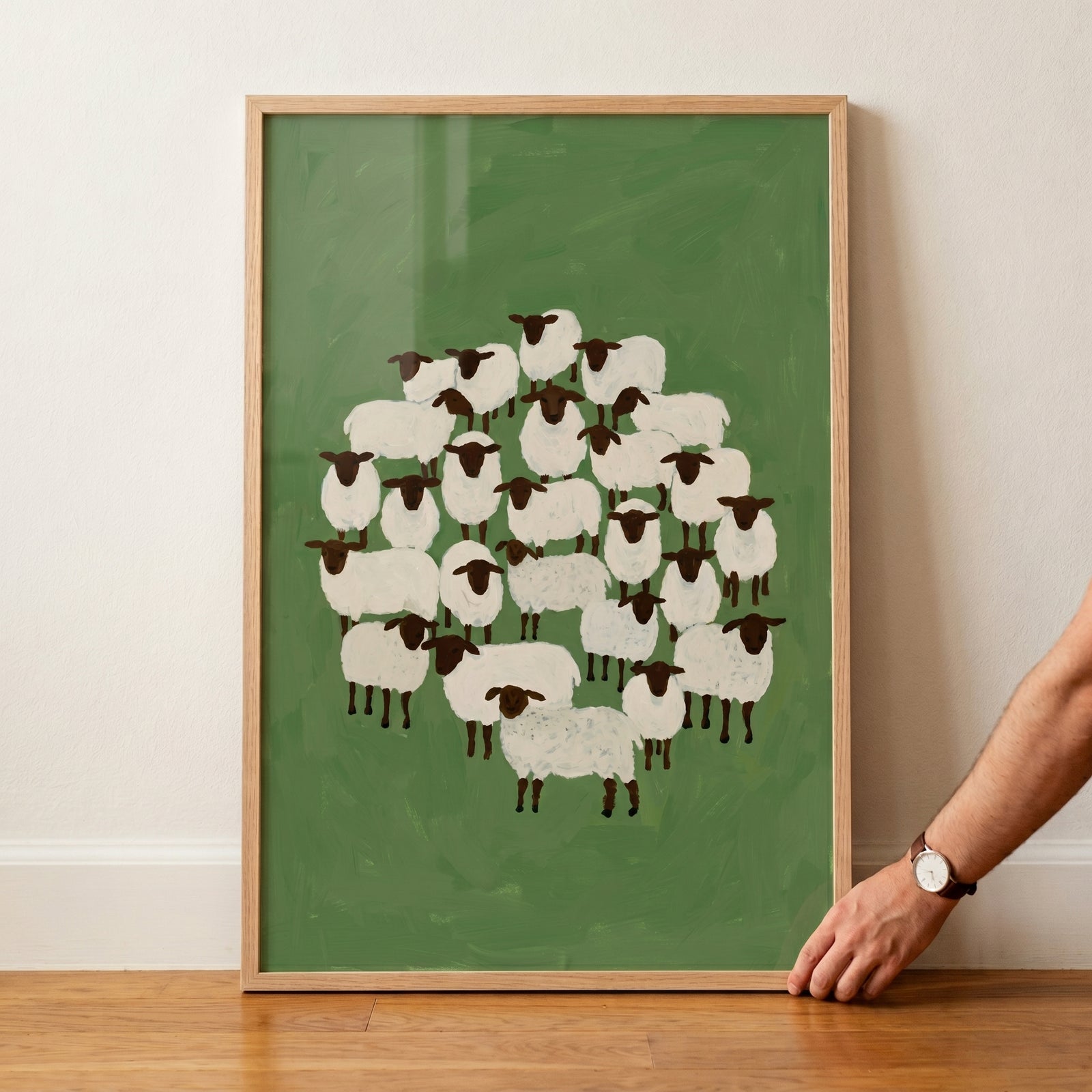 Minimalist black face sheep wall art shows a flock on a green background, held by a hand in a light wood frame.