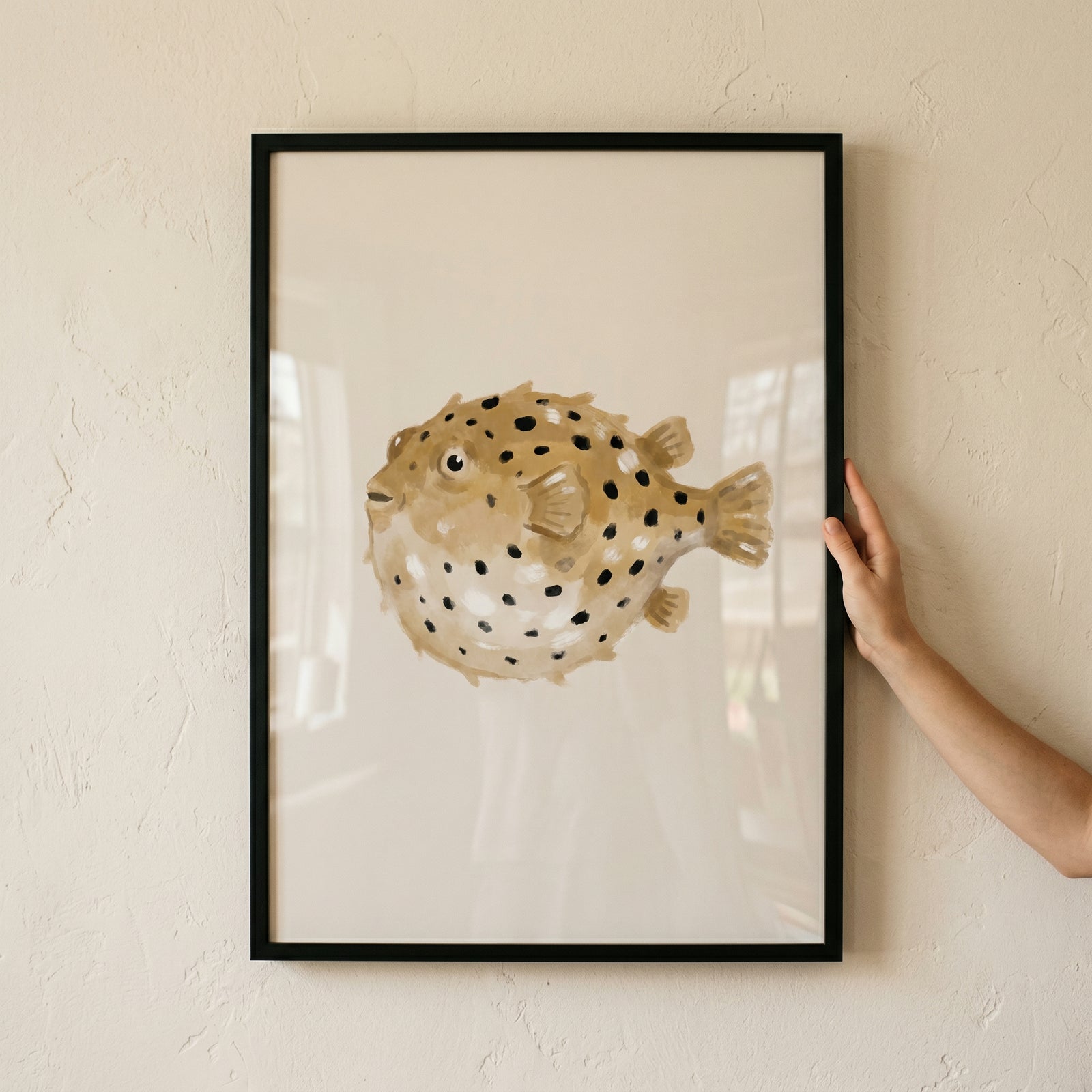 Minimalist black spotted pufferfish wall art in a black frame held by a hand against a textured wall.