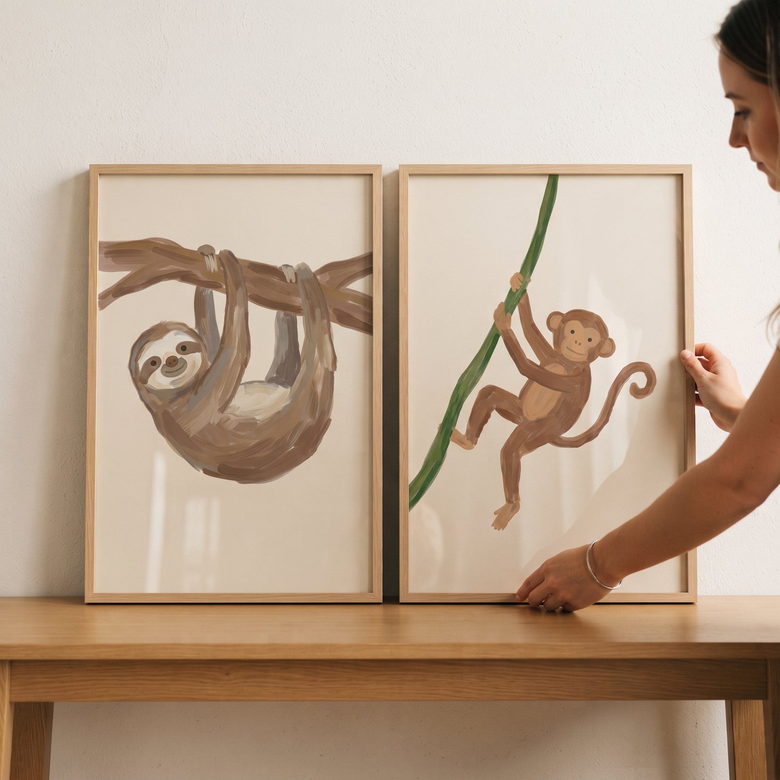 A woman displays a minimalist brown jungle animals wall art set of 2 featuring a sloth and monkey on a table.