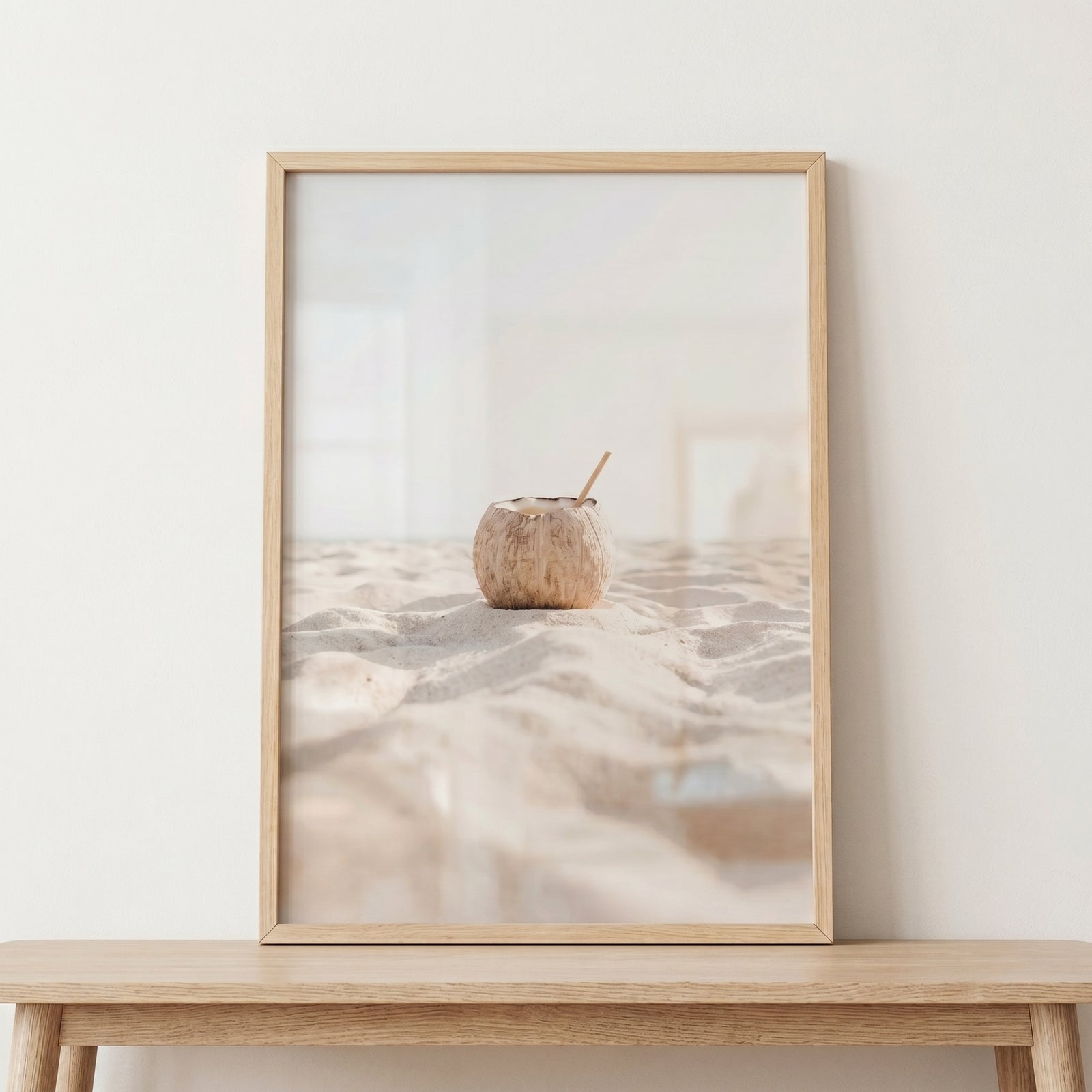 Minimalist creamy white coconut wall art shows a coconut with a straw on sand in a wood frame on a table.