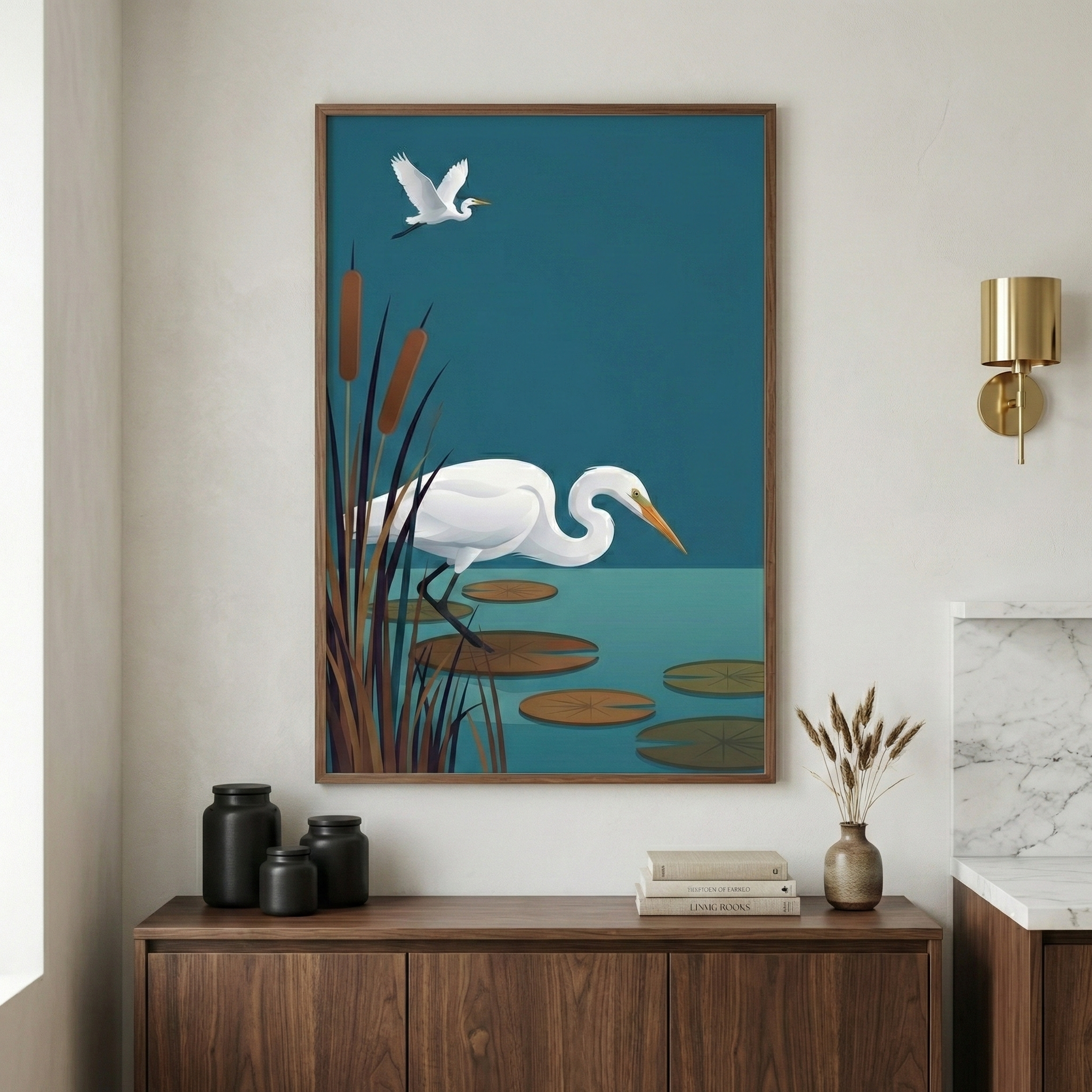 A vertical framed illustration hangs on a neutral-toned wall above a light wooden desk. The artwork shows a minimalist scene of a large white egret standing on a lily pad in a teal-blue pond with tall brown cattails on the left. A smaller egret flies in the upper left corner. On the desk below, there is a ceramic cup, a small notebook, and a pen. A hand in a beige sleeve reaches for a small object on the desk. Bright light enters from a window on the left.