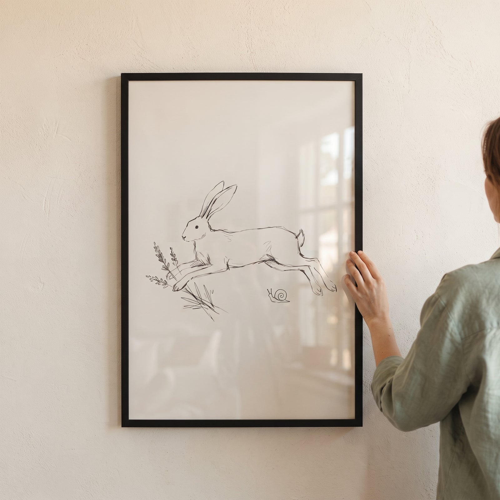 A person holds minimalist leaping bunny wall art in a black frame against a neutral wall.