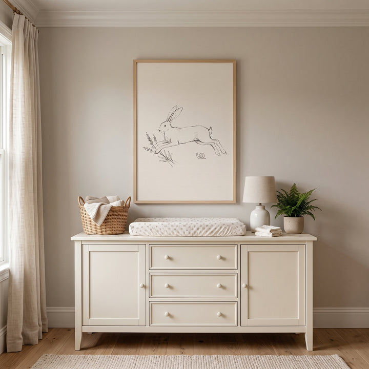 Minimalist leaping bunny wall art hangs above a cream dresser with a changing pad, wicker basket, and small plant.