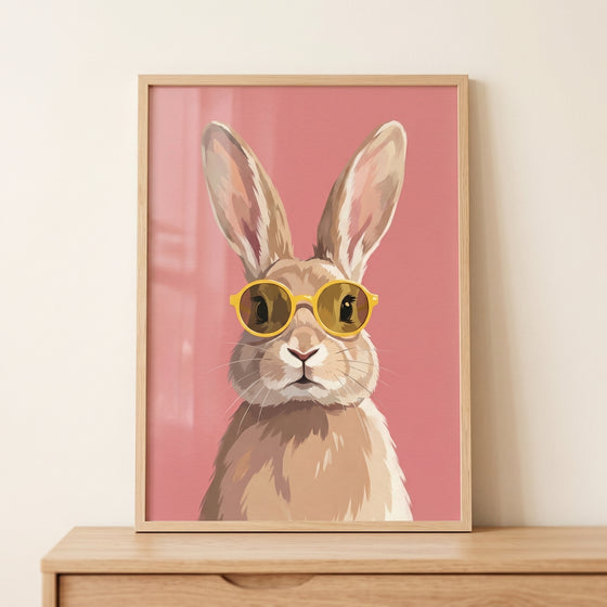 Minimalist pink sunglasses bunny wall art shows a rabbit in yellow shades on a pink background in a light wood frame.