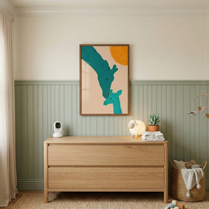 Framed illustration of a tall giraffe bending down to touch noses with a baby giraffe hangs above a wooden dresser in a soft green nursery. The gentle interaction and warm tones add a calming and nurturing focal point to the space.