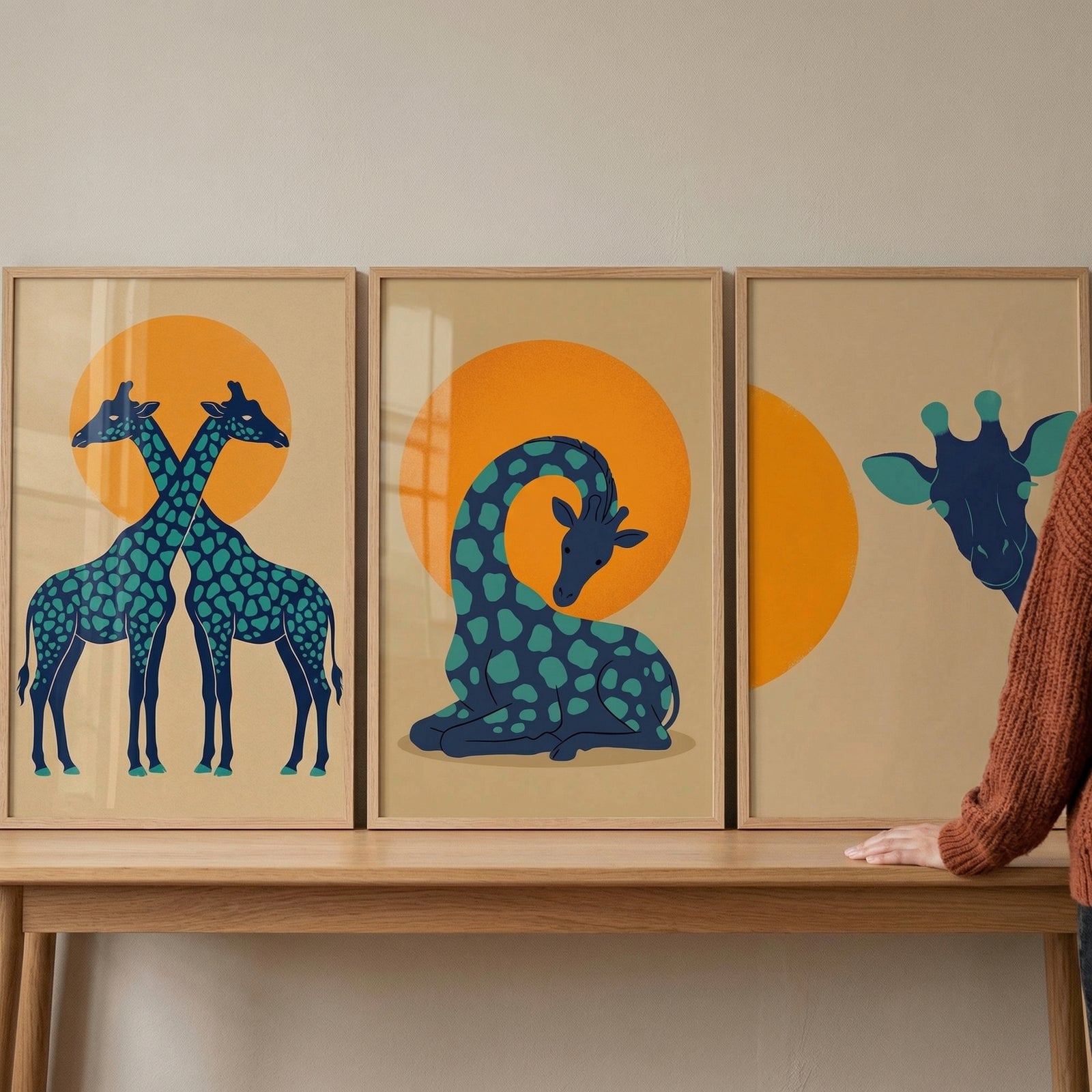 A set of three framed minimalist art pieces sits on a light-colored wooden table against a neutral wall. The art features stylized blue giraffes with teal spots on a light tan background, each centered around a large, bright orange sun. 
