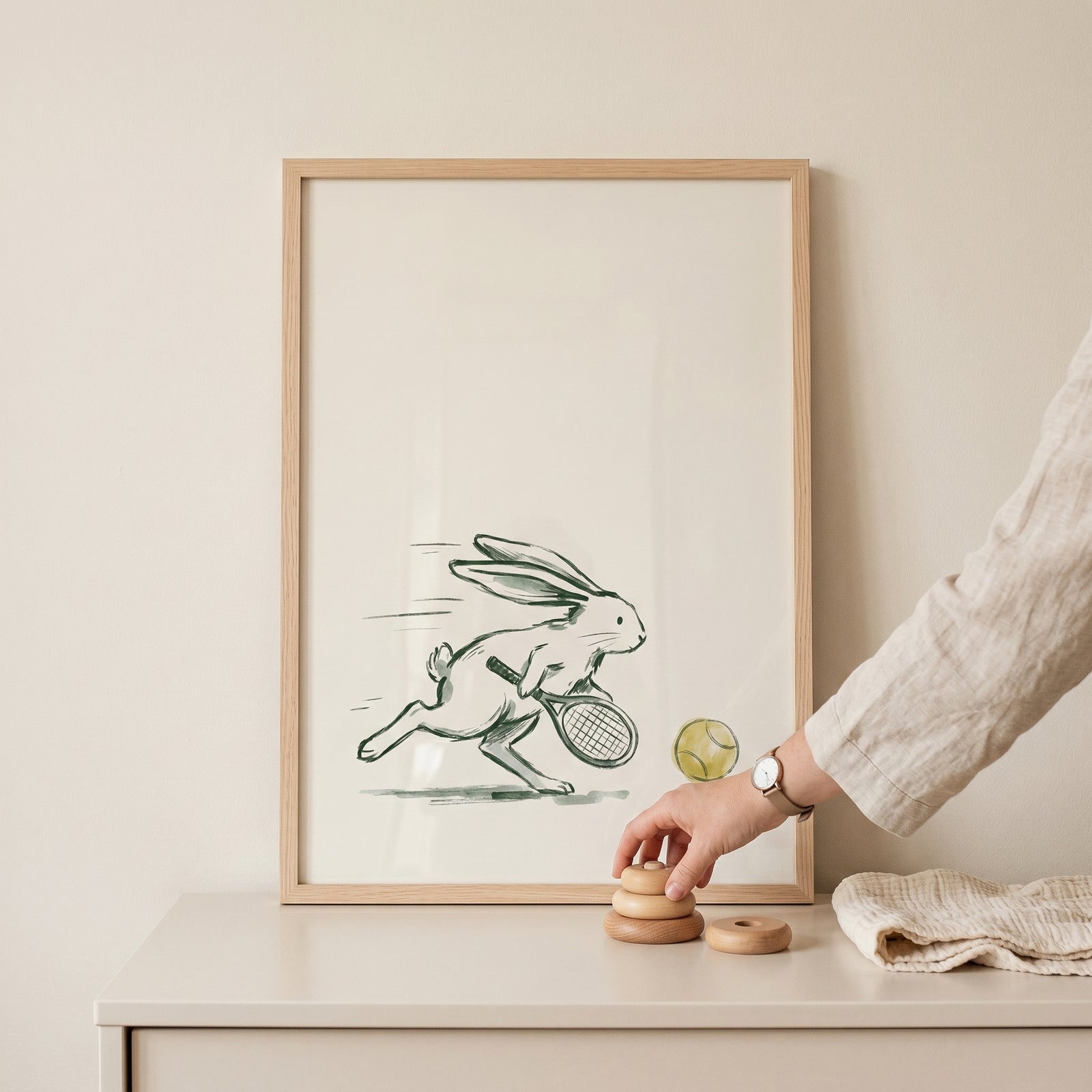 Minimalist tennis rabbit wall art in a wood frame sits on a table next to a person's hand and wooden stacking toys.